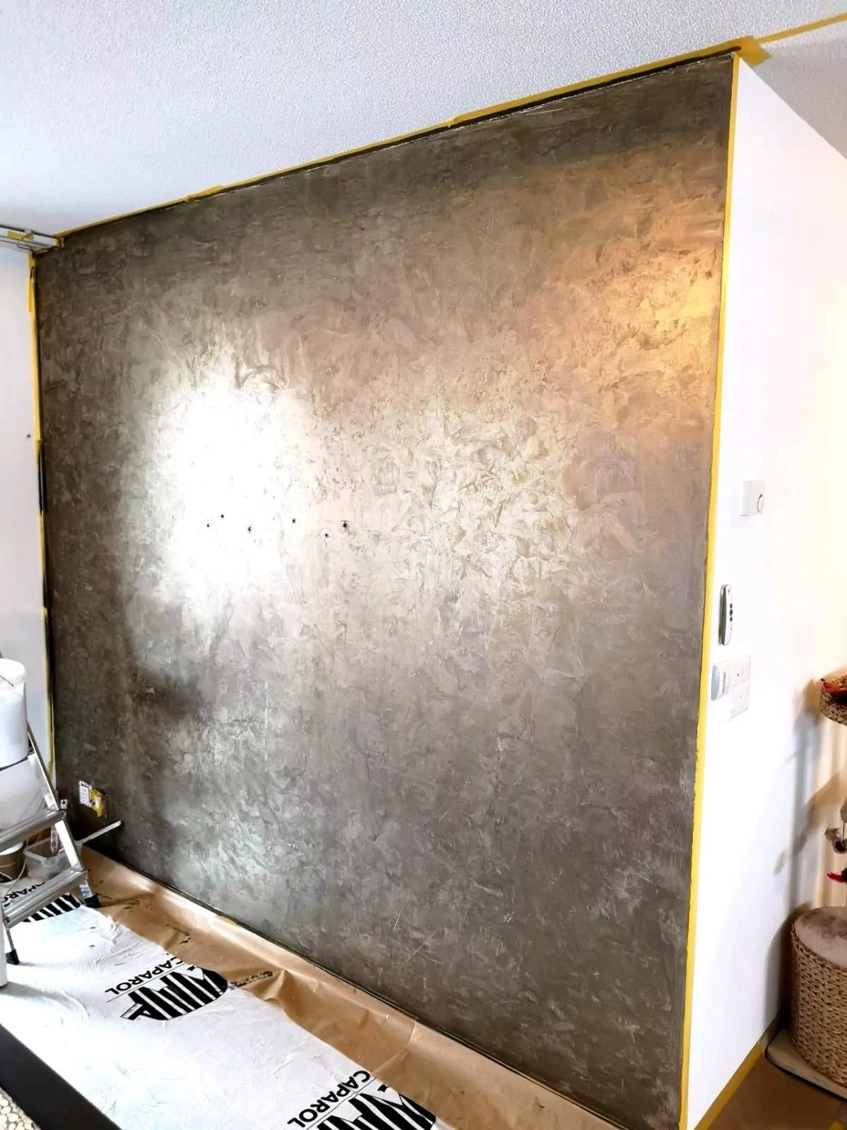 Eine strukturierte Wand mit metallischem Finish in einem Raum, eingefasst mit gelbem Klebeband. Der Boden ist mit Schutzmaterial mit der Aufschrift „GRIPSET“ bedeckt. Gegenstände wie ein Stuhl und ein Behälter sind teilweise in der Nähe der Wand sichtbar.