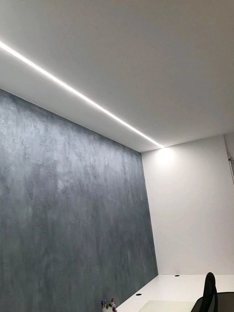 Ein Raum mit minimalistischem Design mit einer grau strukturierten Akzentwand, einer eleganten weißen Decke mit eingelassener linearer Beleuchtung und einem teilweise sichtbaren Schreibtisch in der unteren Ecke mit Stühlen und einigen Büromaterialien.