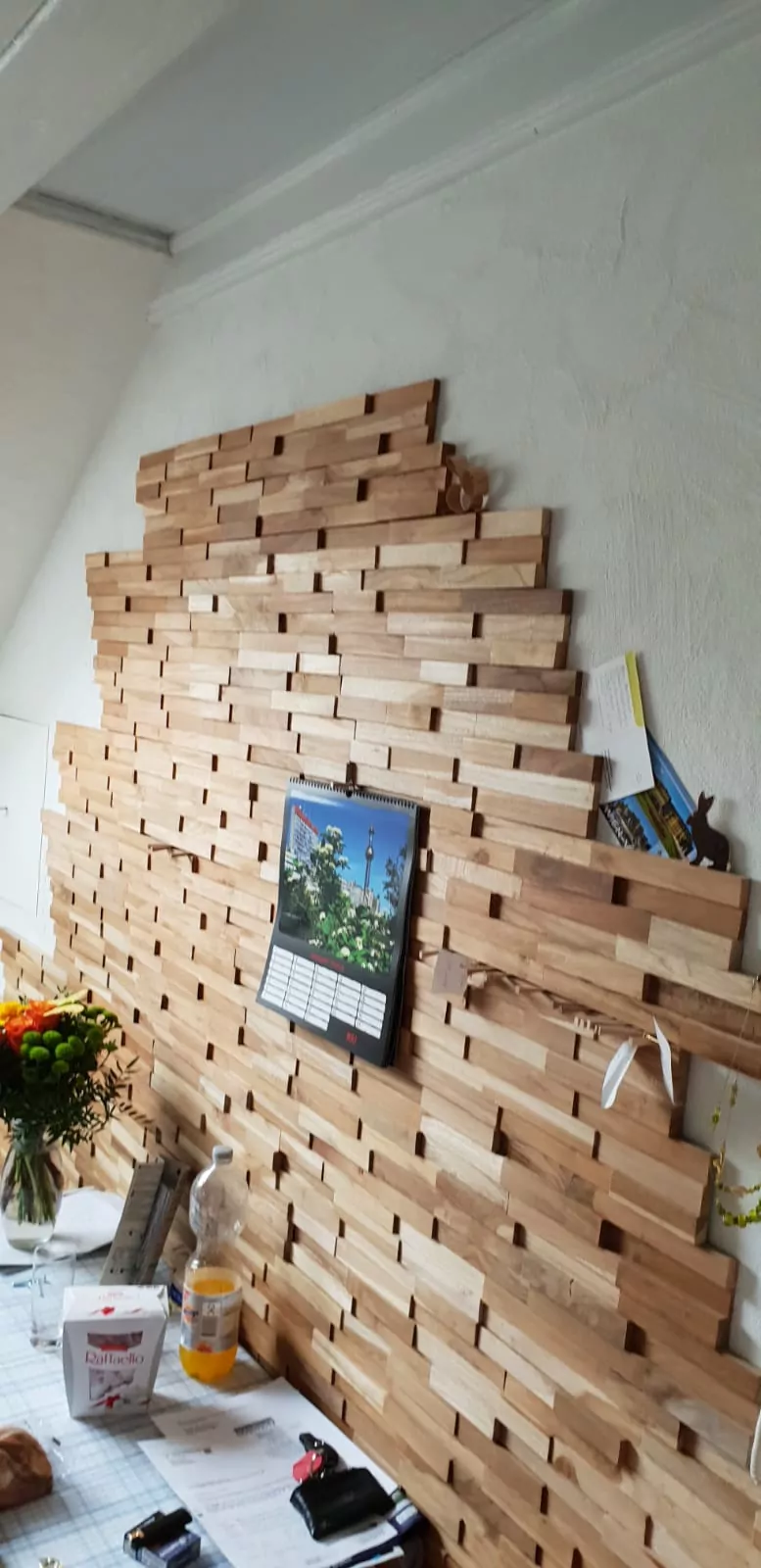 Es wird Holzwandkunst aus ineinander greifenden rechteckigen Blöcken ausgestellt. An der Wand hängt ein Kalender. In der Nähe steht auf einem Tisch eine Flasche, Papiere und ein Blumenstrauß.