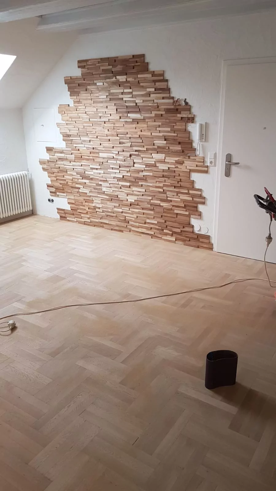 Ein Raum mit Holzparkettboden und einer Akzentwand, die teilweise mit versetzten Holzpaneelen verkleidet ist. Auf dem Boden liegen ein Stromkabel und ein kleiner Eimer. Der Raum hat eine weiße Wand, eine Tür und einen Heizkörper unter einem Fenster.