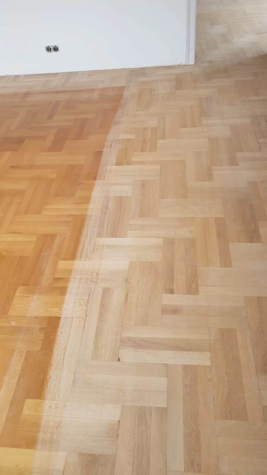 Ein Holzparkettboden mit einer klaren Abgrenzung in zwei Bereiche: Die linke Seite ist dunkler und unbehandelt, während die rechte Seite heller ist und frisch abgeschliffen oder behandelt erscheint. Im Hintergrund sind an der Wand zwei Steckdosen zu sehen.