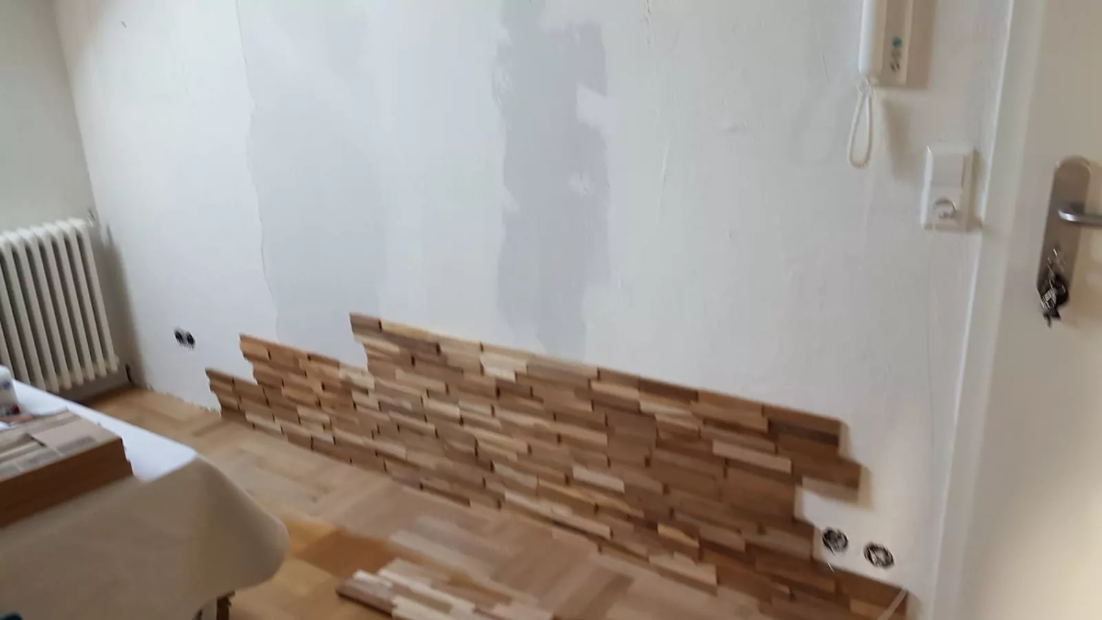 Teilweise angebrachte Holzvertäfelung an einer Wohnzimmerwand, wobei einige Bretter fehlen. Links ist ein Heizkörper zu sehen, ein Tisch ist teilweise sichtbar. Rechts neben einer geschlossenen Tür ist eine Gegensprechanlage angebracht.