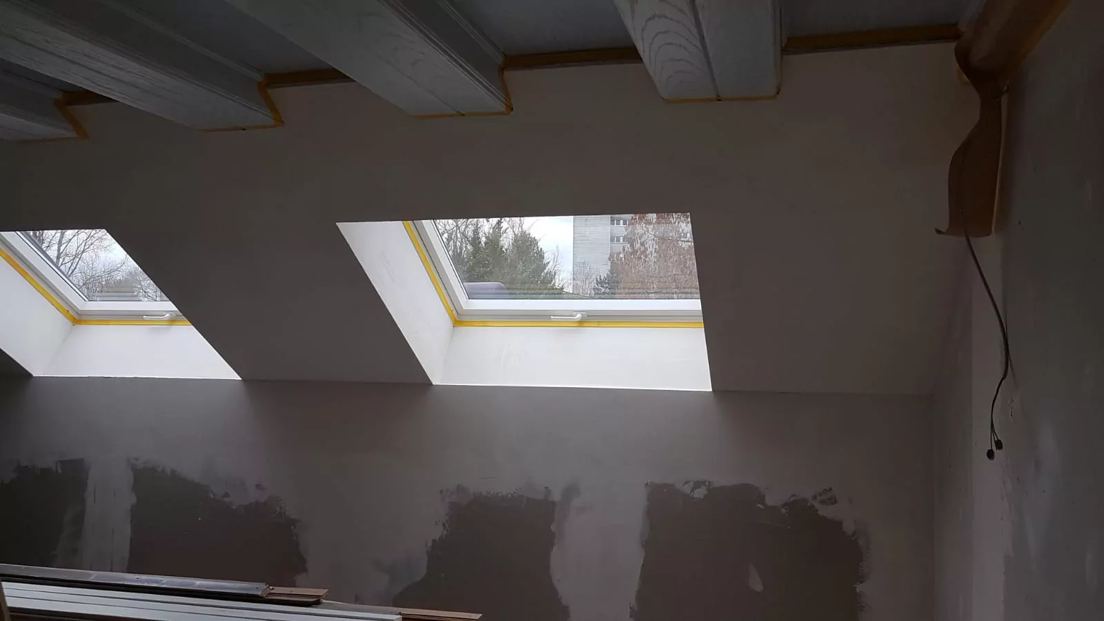 Ein Raum mit unfertigen Wänden und freiliegenden Holzbalken an der Decke verfügt über zwei Oberlichtfenster. Die Fenster bieten einen Blick auf Bäume und ein Gebäude draußen. An der rechten Seite der Decke hängt ein Kabel.