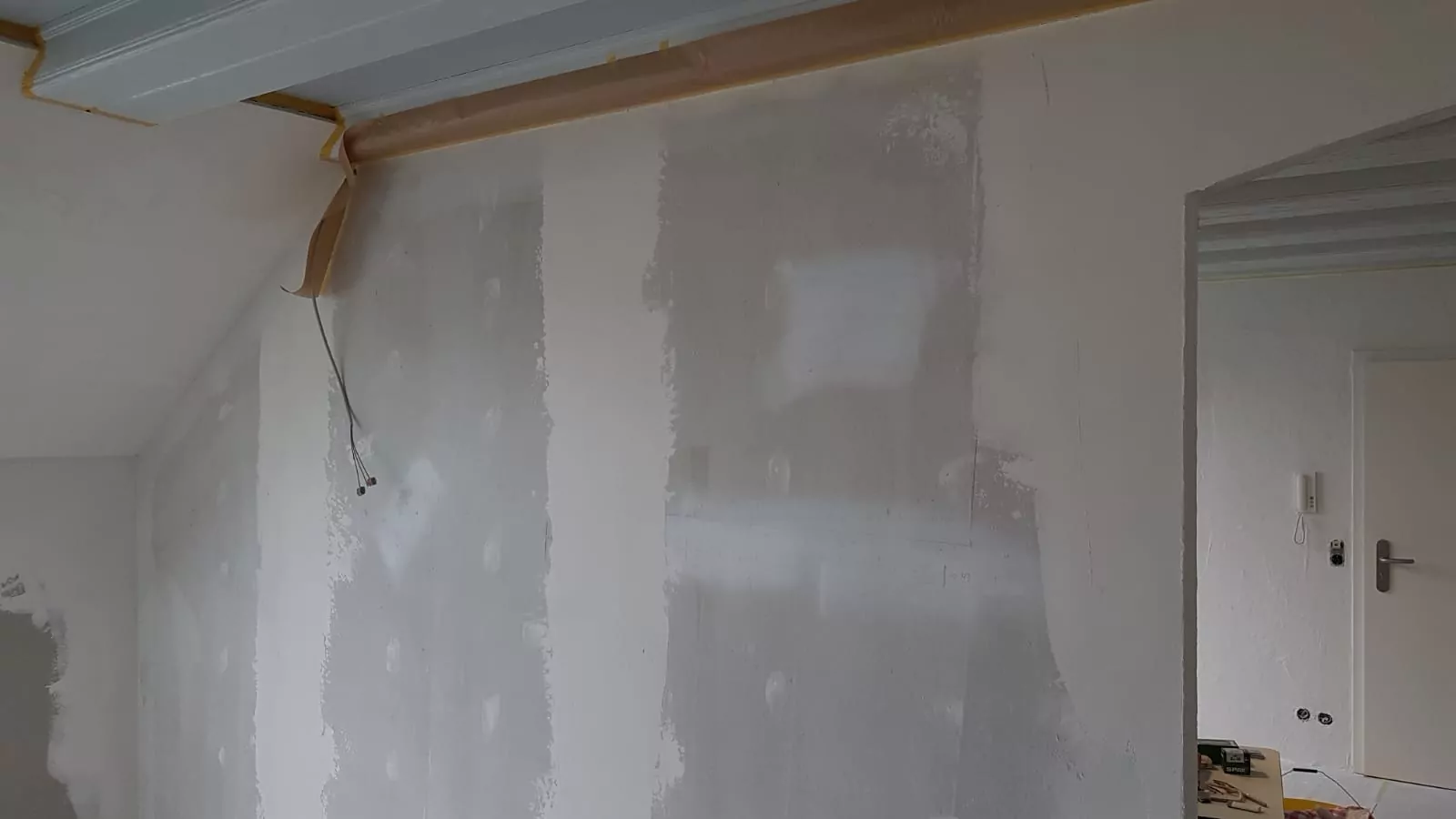 Ein teilweise renovierter Raum mit Wänden, die Putzstellen und freiliegende Gipskartonplatten aufweisen. Die Decke und Teile der Wände sind mit Schutzklebeband abgedeckt. Auf der rechten Seite befindet sich eine Tür mit einem Metallgriff. Von der Decke hängt ein herabhängendes Kabel.