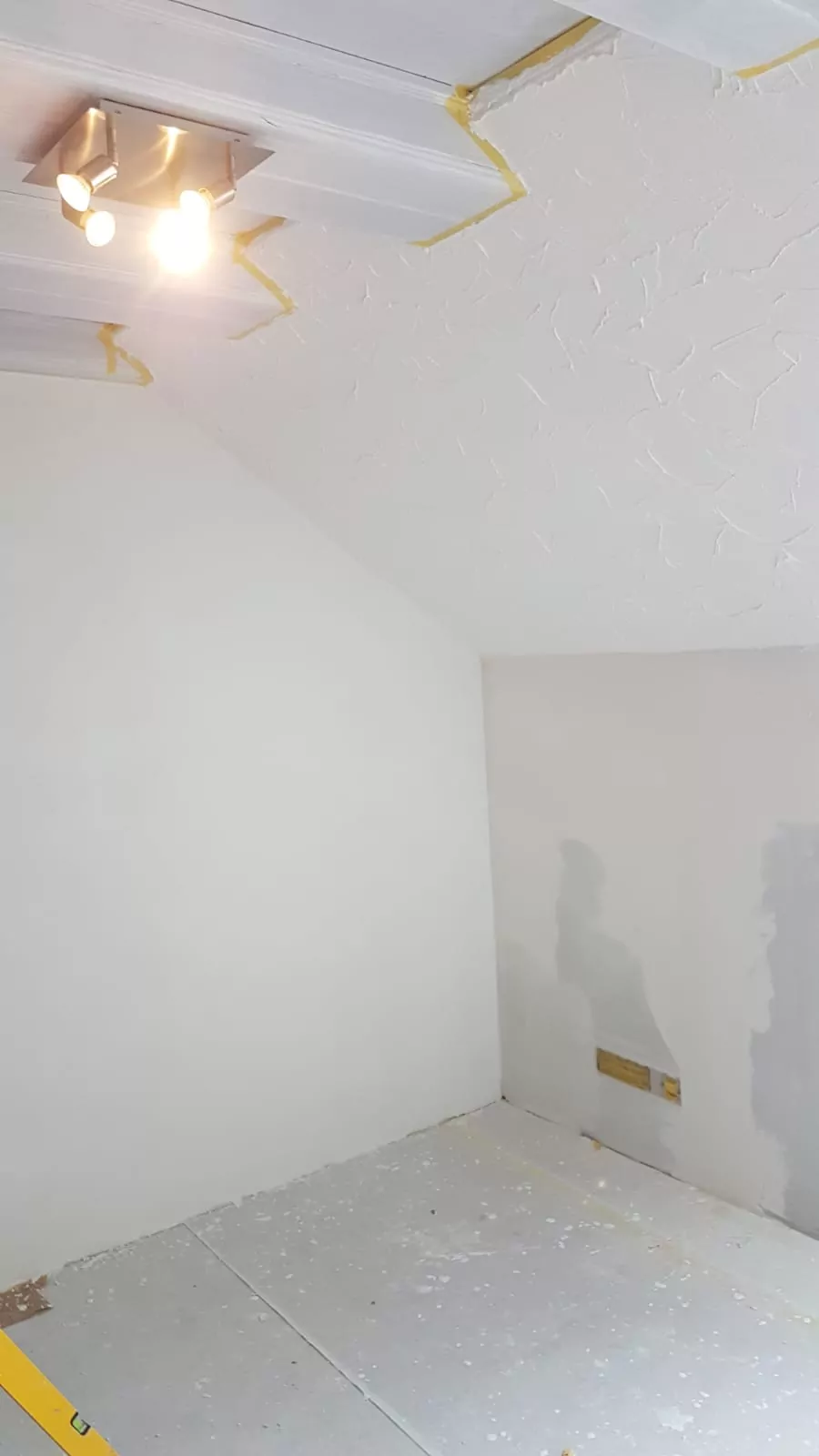 Ein Raum mit unfertigen Wänden und Decken, die teilweise eine strukturierte Oberfläche aufweisen. An der Decke befindet sich ein Spotlicht, und der Boden ist mit Schutzmaterial bedeckt. An den Rändern der Decke sind abgeklebte Bereiche zu sehen, was auf laufende Renovierungsarbeiten hindeutet.