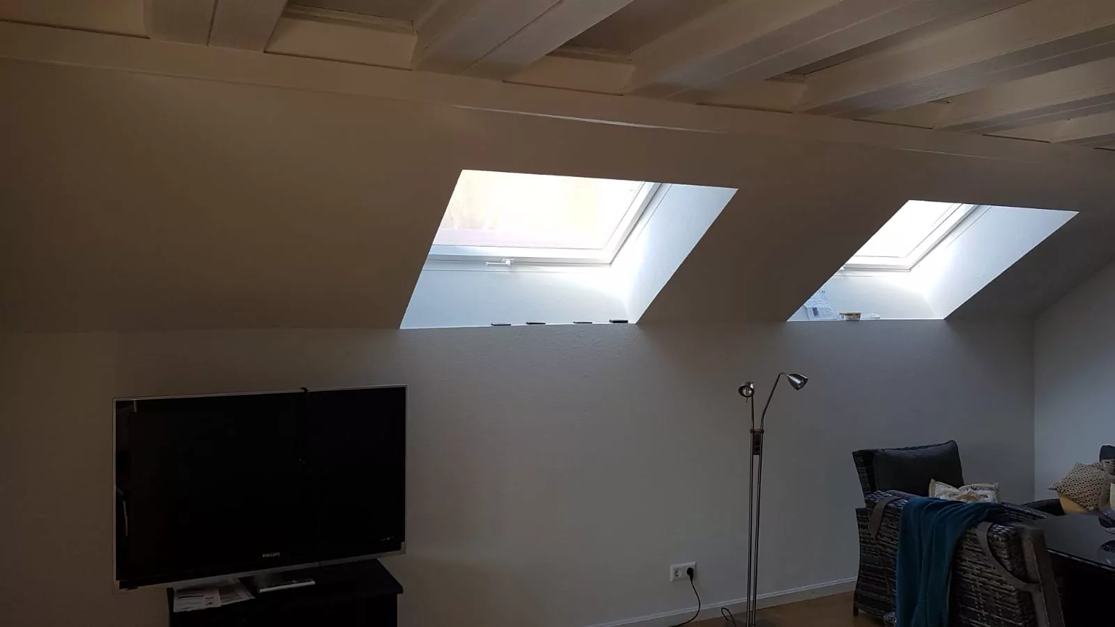 Ein Raum mit Dachschrägen, der durch drei Oberlichter Tageslicht hereinlässt. Darunter stehen ein Fernseher auf einem Ständer und eine Stehlampe. Auf der rechten Seite sind einige Sitzgelegenheiten mit Kissen und einer blauen Decke zu sehen. Die Wände sind hell.