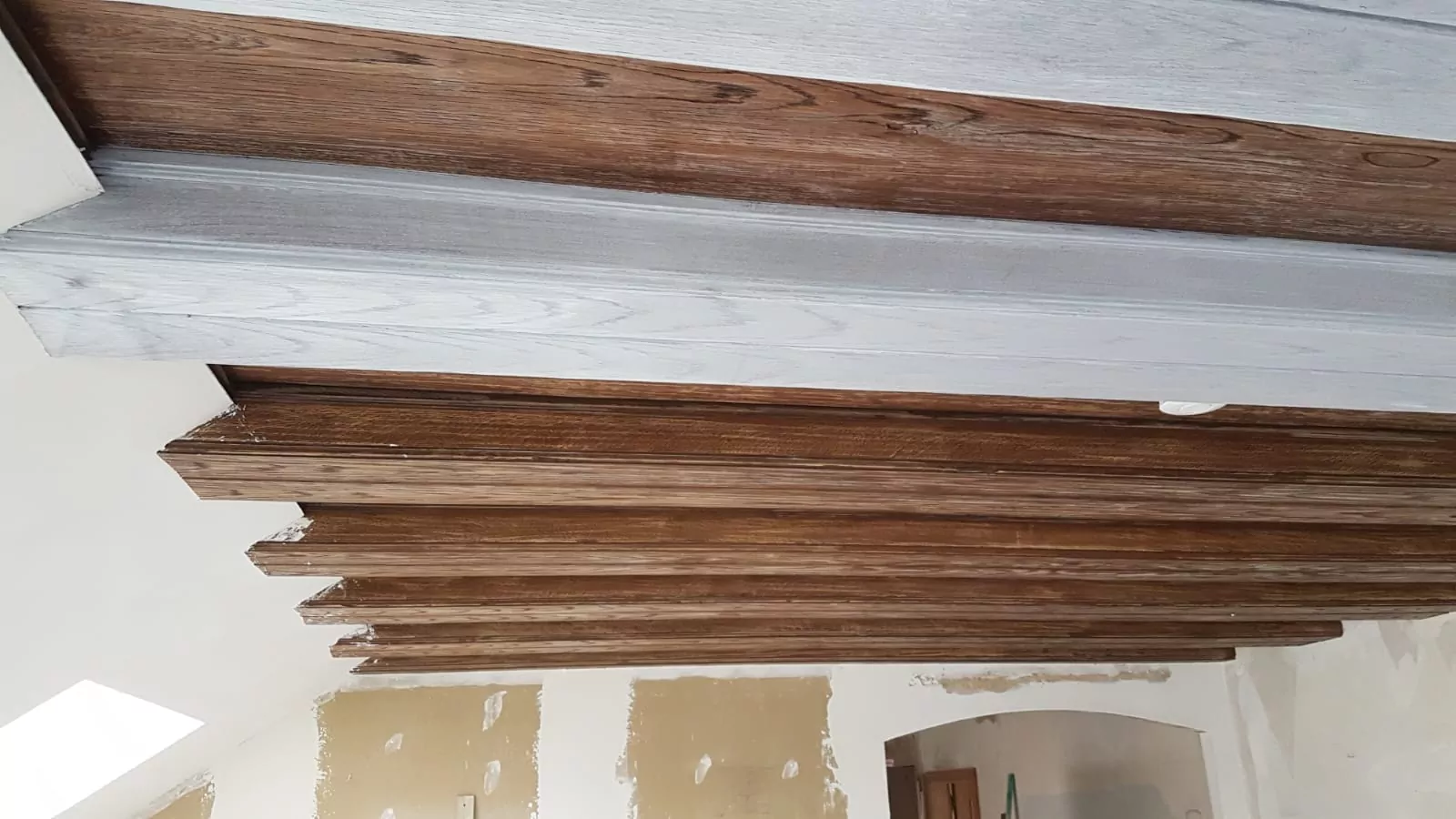 An der Decke sind Holzbalken angebracht. Die Balken wechseln zwischen dunklen und hellen Holztönen. Der Raum ist teilweise verputzt und zeigt einige unfertige Wände mit Flicken. In der oberen linken Ecke befindet sich ein kleines Fenster.