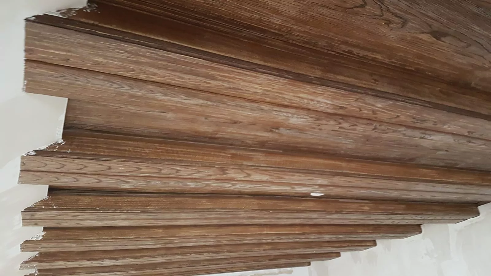 Eine Reihe von Holzbrettern bildet ein versetztes Deckenelement mit einer natürlichen Holzmaserung. Die Bretter sind parallel angeordnet und bilden ein lineares Muster über die Decke. Die umgebenden Wände sind unbemalt.