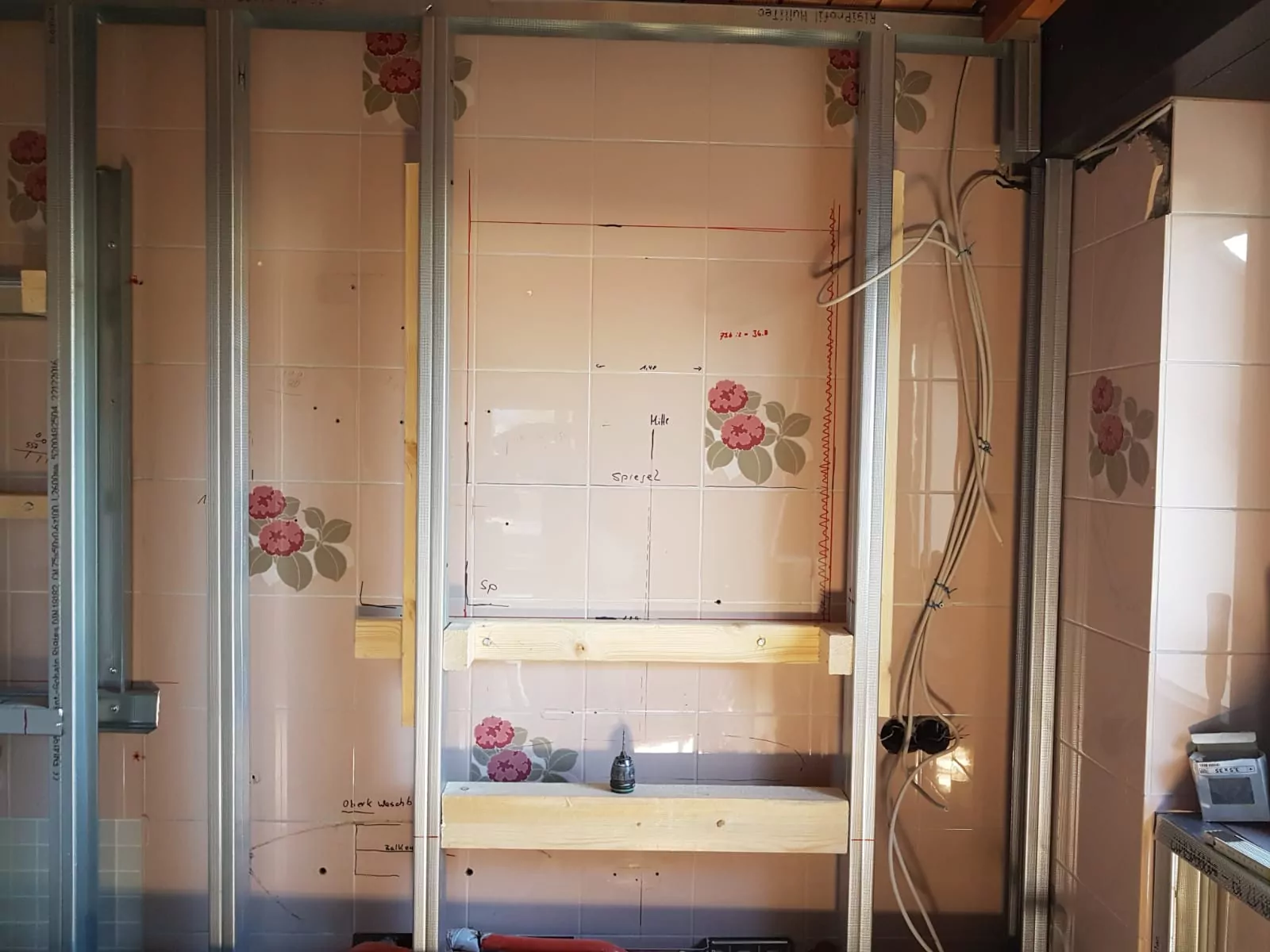 Eine teilweise fertiggestellte Wand zeigt Metallrahmen und Holzstützen vor Fliesen mit Blumenmuster. Aus der Wand ragen Kabel hervor, die auf laufende Elektroarbeiten hinweisen.