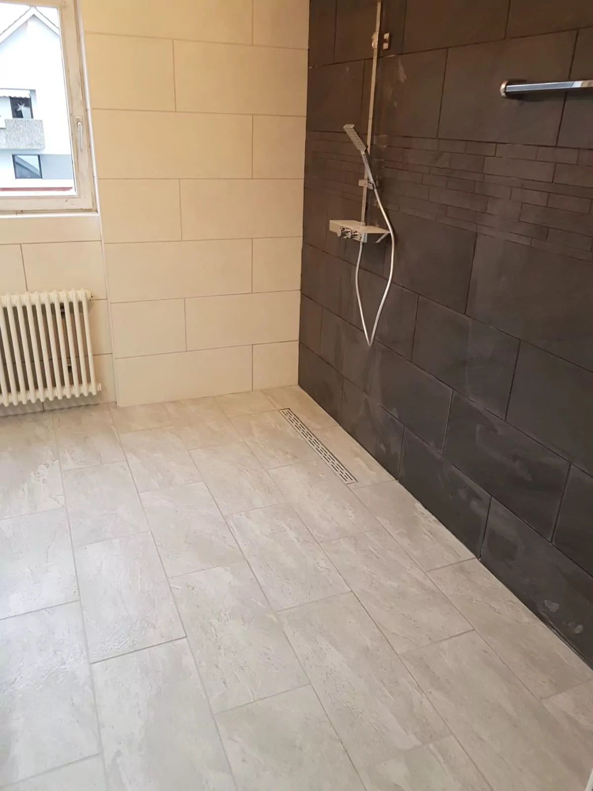 Ein Badezimmer verfügt über eine ebenerdige Dusche mit schwarzen Fliesen und einem modernen Handbrausenkopf. Der Boden ist mit hellgrauen Fliesen und einem linearen Abfluss bedeckt. Auf der linken Seite ist ein Fenster mit einem Heizkörper darunter zu sehen, der für natürliches Licht sorgt.