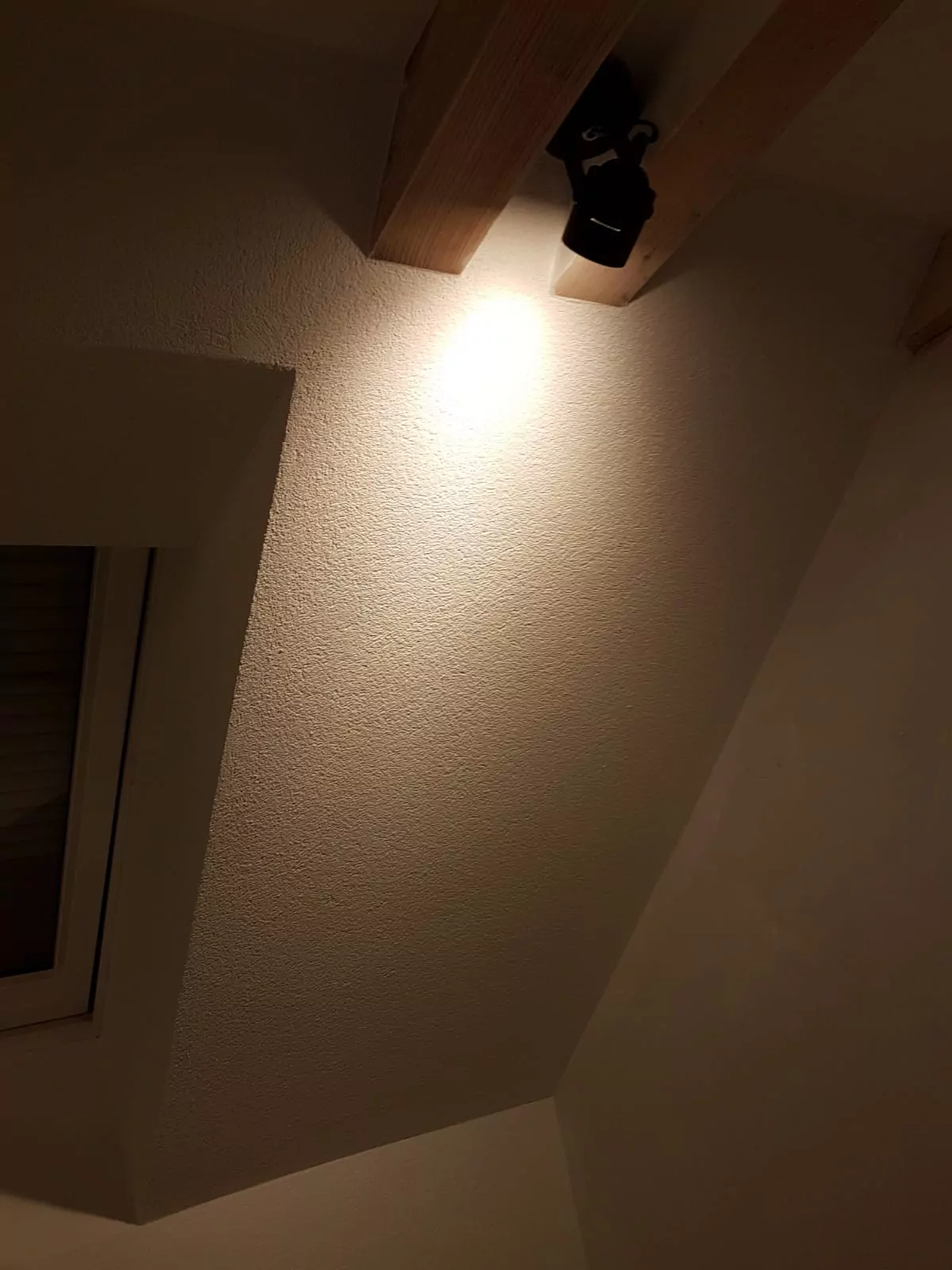 Eine Zimmerdecke, die teilweise von einem Scheinwerfer beleuchtet wird. Holzbalken sind sichtbar und auf der linken Seite des Bildes ist ein Fenster teilweise zu sehen.