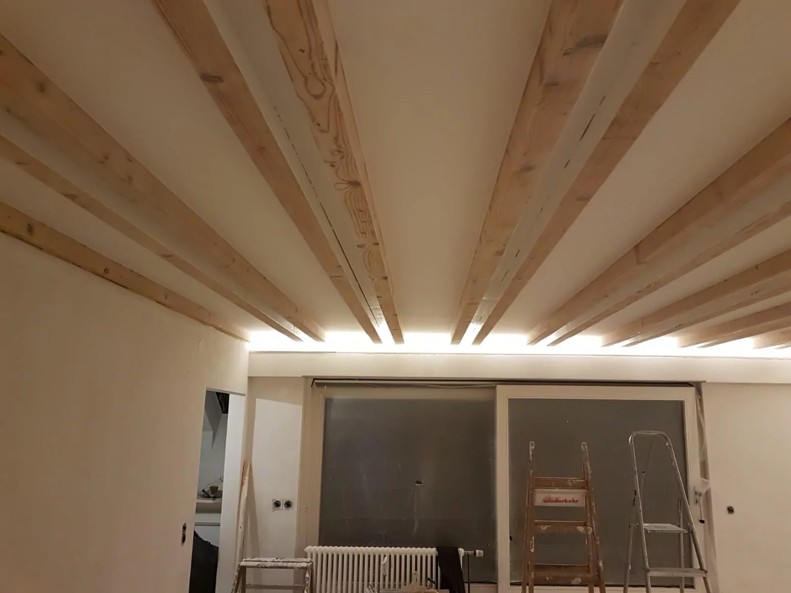 Ein im Bau befindlicher Raum mit Holzbalken an der Decke. Es sind Leitern und ein Heizkörper vorhanden und an einer Wand ist ein großes rahmenloses Fenster oder eine Glastür sichtbar.