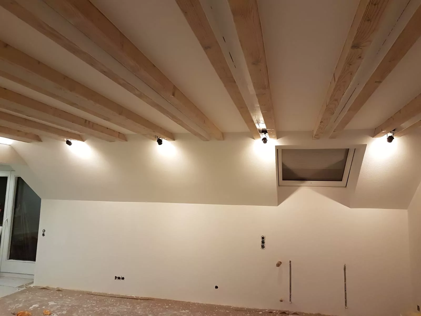 Ein im Bau befindlicher Raum mit freiliegenden Holzbalken an der Decke. Die Wände sind teilweise unfertig, Armaturen und Steckdosen sind sichtbar. Im oberen Teil der Wand ist ein Fenster mit Jalousie eingelassen. Die Böden scheinen nicht mit Teppich ausgelegt und ungeschliffen zu sein.