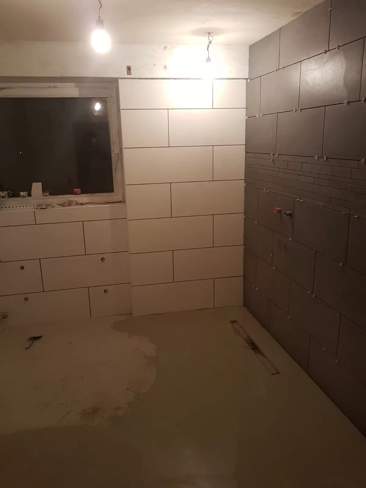 Ein teilweise gefliester Raum im Bau, mit weißen Fliesen an einer Wand und dunkelgrauen Fliesen an einer anderen. An der linken Wand befindet sich ein Fenster. Der Boden ist mit einer glatten Betonschicht bedeckt. Zwei hängende Glühbirnen beleuchten den Raum.