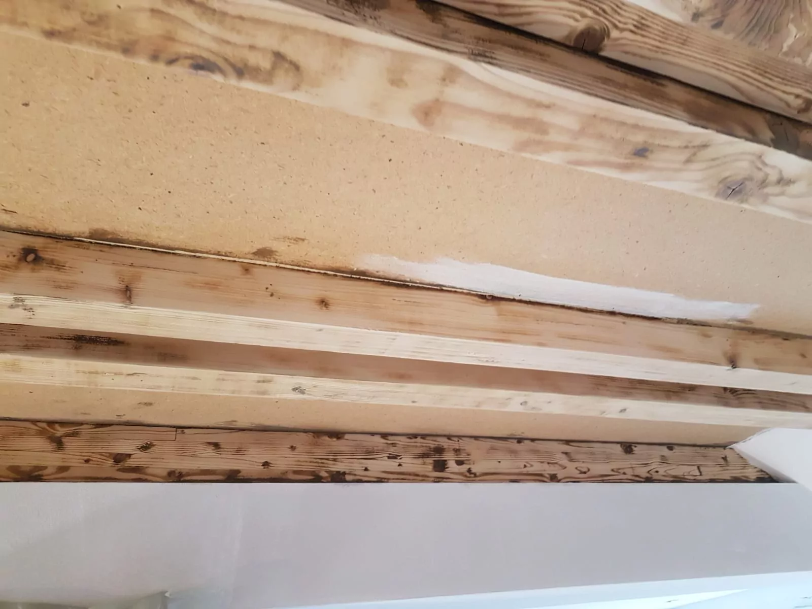 Das Bild zeigt eine Decke mit freiliegenden Holzbalken. Einige Balken weisen dunkle Flecken oder Brandflecken auf, andere sind unbehandelt oder teilweise gestrichen. An den Rändern ist ein kleiner Abschnitt Gipskartonplatten sichtbar.