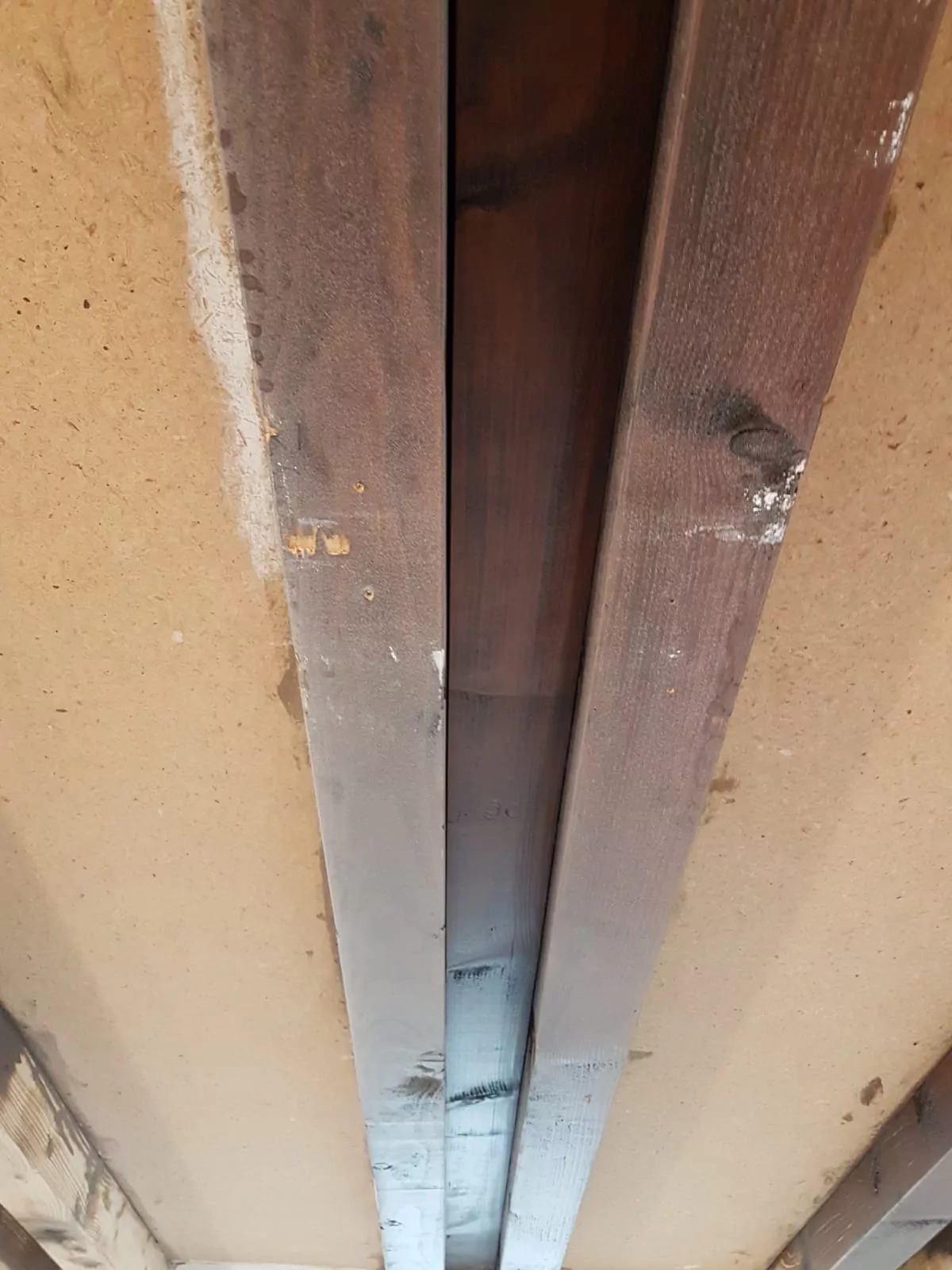 Nahaufnahme einer Decke mit freiliegenden Holzbalken und einer Spanplatte dazwischen. Das Holz weist Flecken auf, auf der Oberfläche sind einige Flecken und Schmierer zu sehen. Die Balken sind parallel und gleichmäßig über die Decke verteilt.