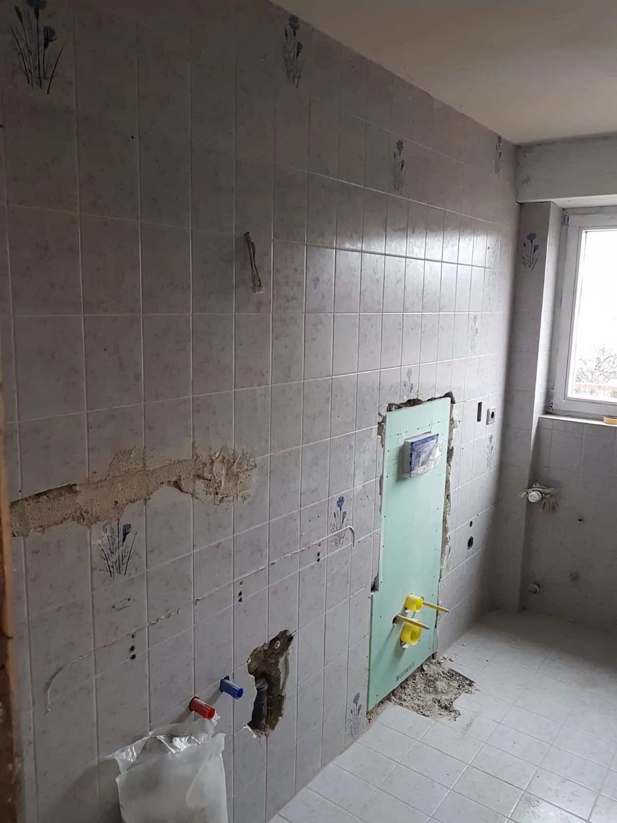 Ein teilweise renoviertes Badezimmer mit entfernten Armaturen. Die Wände sind mit hellgrauen Fliesen mit blauem Blumenmuster bedeckt, einige Fliesenabschnitte fehlen. Sanitärarmaturen und ein Fenster sind sichtbar. Der Boden hat passende Fliesen.