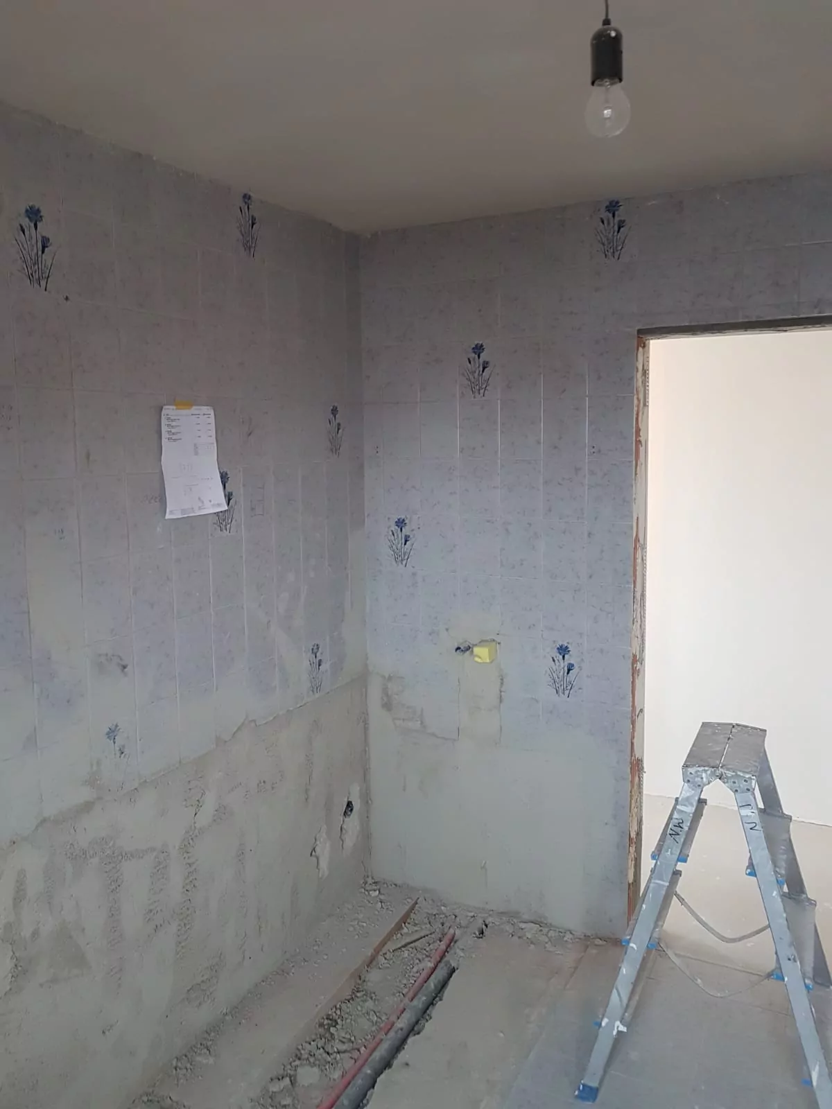 Ein teilweise renovierter Raum mit kahlen Wänden, die teilweise mit Fliesen mit einem einfachen Blumenmuster bedeckt sind. Neben der Tür steht eine Leiter. Ein Stück Papier ist an die Wand geklebt und Baumaterialien liegen verstreut auf dem Boden.