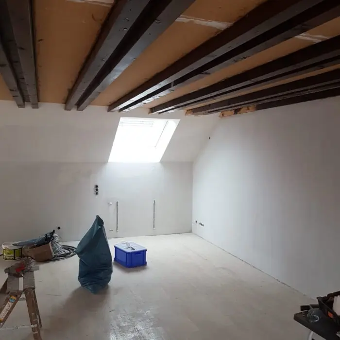 Ein teilweise renovierter Raum mit unfertigen Holzbalken an der Decke und weiß verputzten Wänden. Ein Oberlicht sorgt für natürliches Licht. Auf dem nackten Boden liegen verstreut Gegenstände wie eine Leiter, Farbdosen und eine blaue Aufbewahrungsbox.