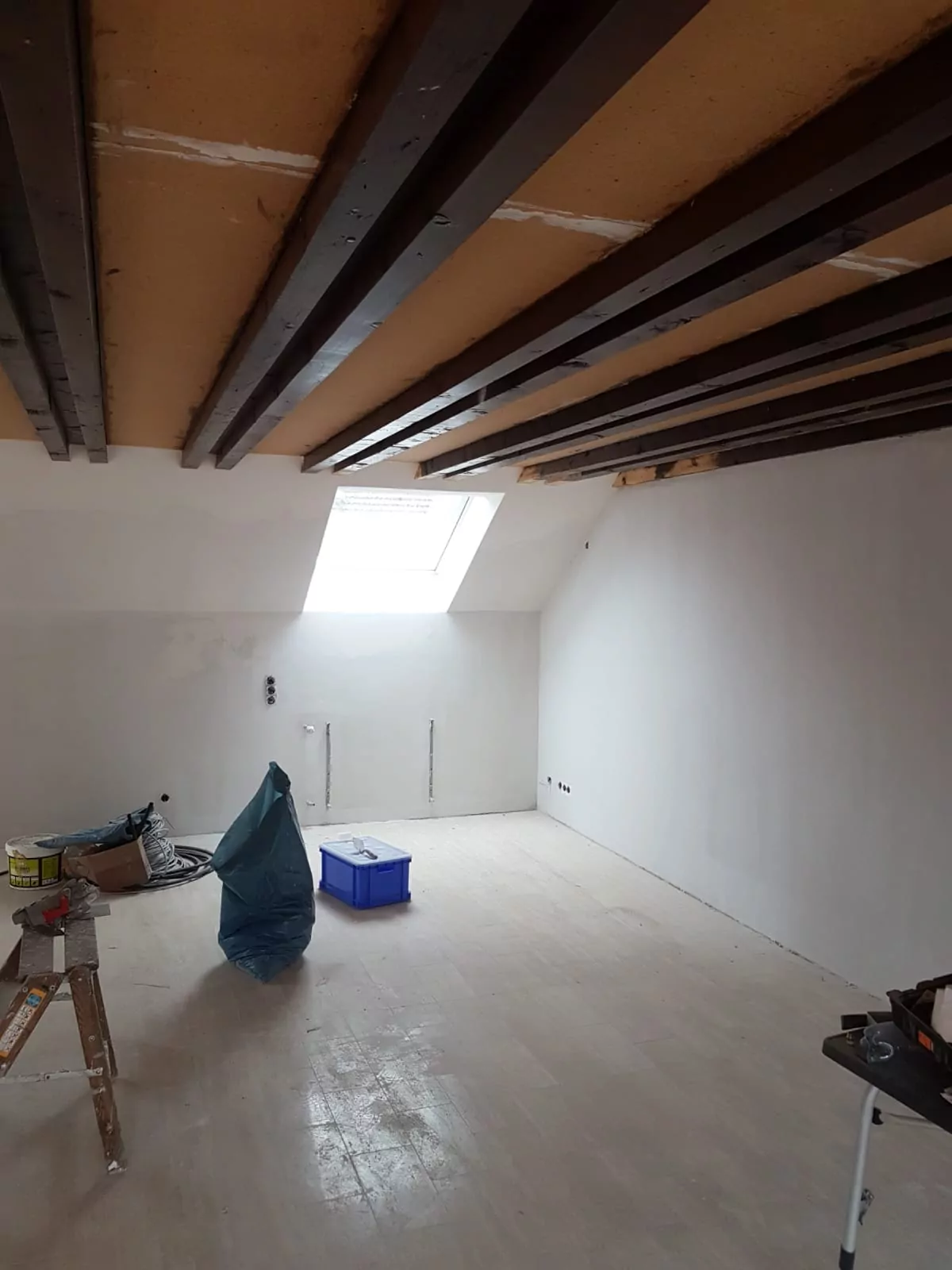 Ein teilweise renovierter Raum mit unfertigen Holzbalken an der Decke und weiß verputzten Wänden. Ein Oberlicht sorgt für natürliches Licht. Auf dem nackten Boden liegen verstreut Gegenstände wie eine Leiter, Farbdosen und eine blaue Aufbewahrungsbox.
