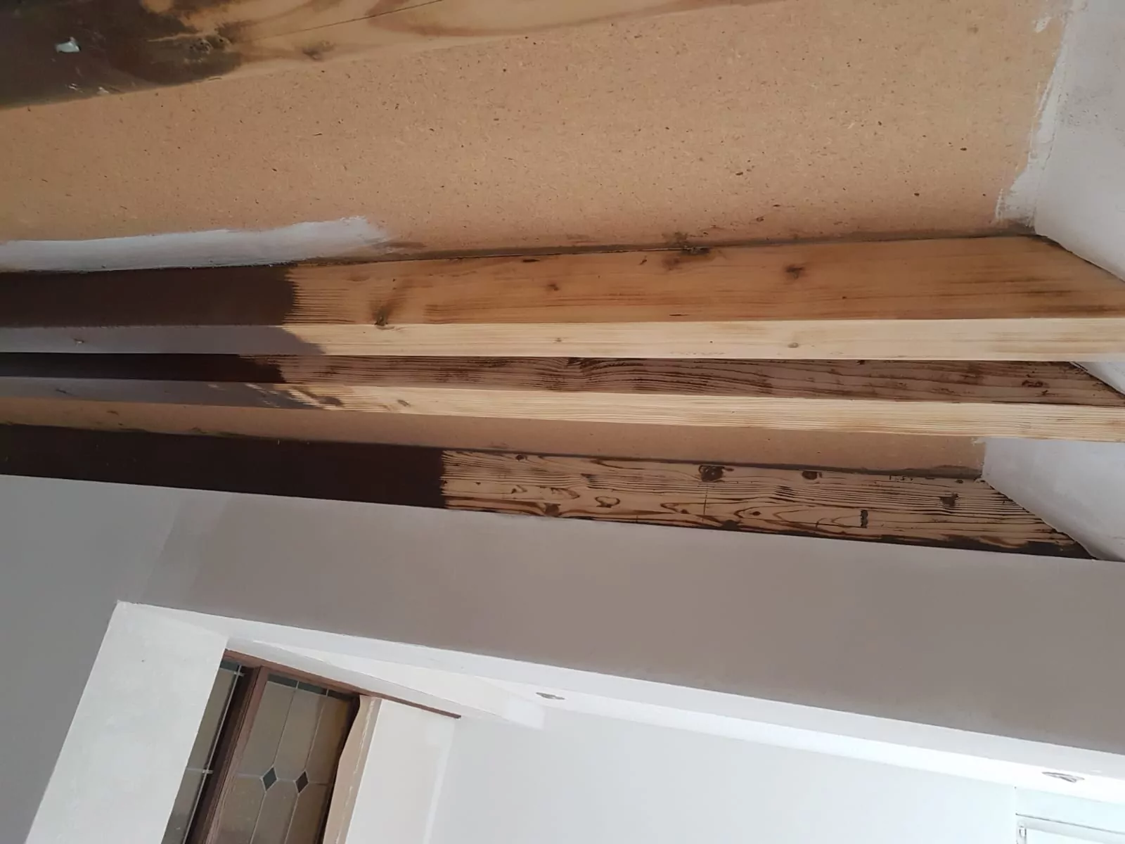 Eine Deckenecke mit freiliegenden Holzbalken in verschiedenen Färbungsstadien. Einige Balken sind teilweise dunkel gebeizt, während andere unbearbeitet bleiben. Die umgebenden Wände sind weiß und beige gestrichen.