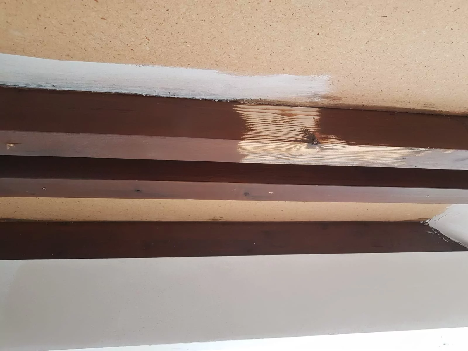 Es sind Holzbalken an der Decke mit sichtbaren Wasserschäden zu sehen. Ein Balken hat einen helleren, verfärbten Fleck, der auf Feuchtigkeitsbelastung hinweist. Die umgebende Decke ist cremefarben und die Balken sind dunkelbraun.