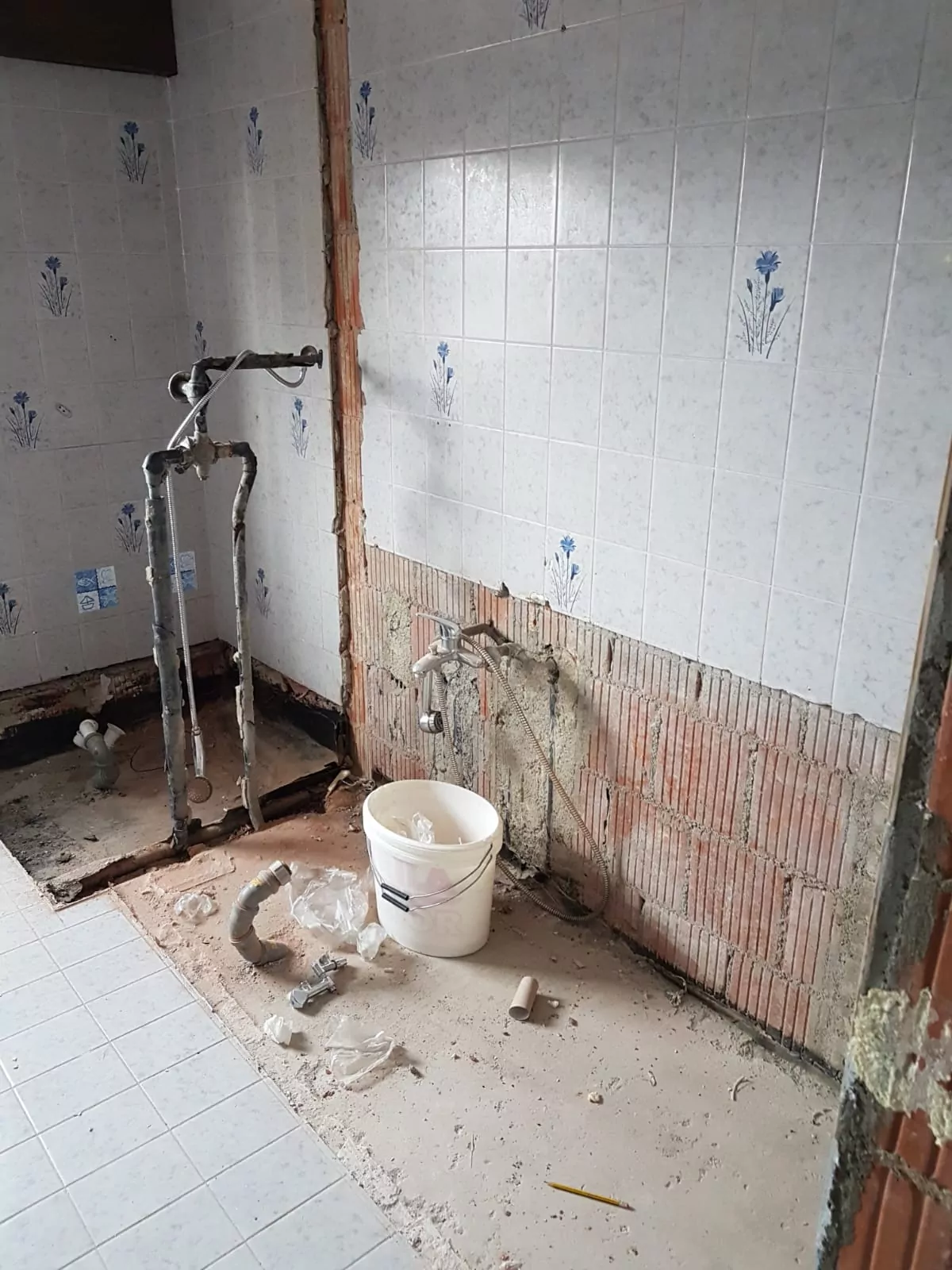 Ein teilweise renoviertes Badezimmer mit freiliegenden Ziegelwänden und Rohrleitungen. Die Keramikfliesen darüber haben ein Blumenmuster. Rohre sind sichtbar und auf dem Fliesenboden liegen Baumaterialien und Schutt, darunter ein weißer Eimer.