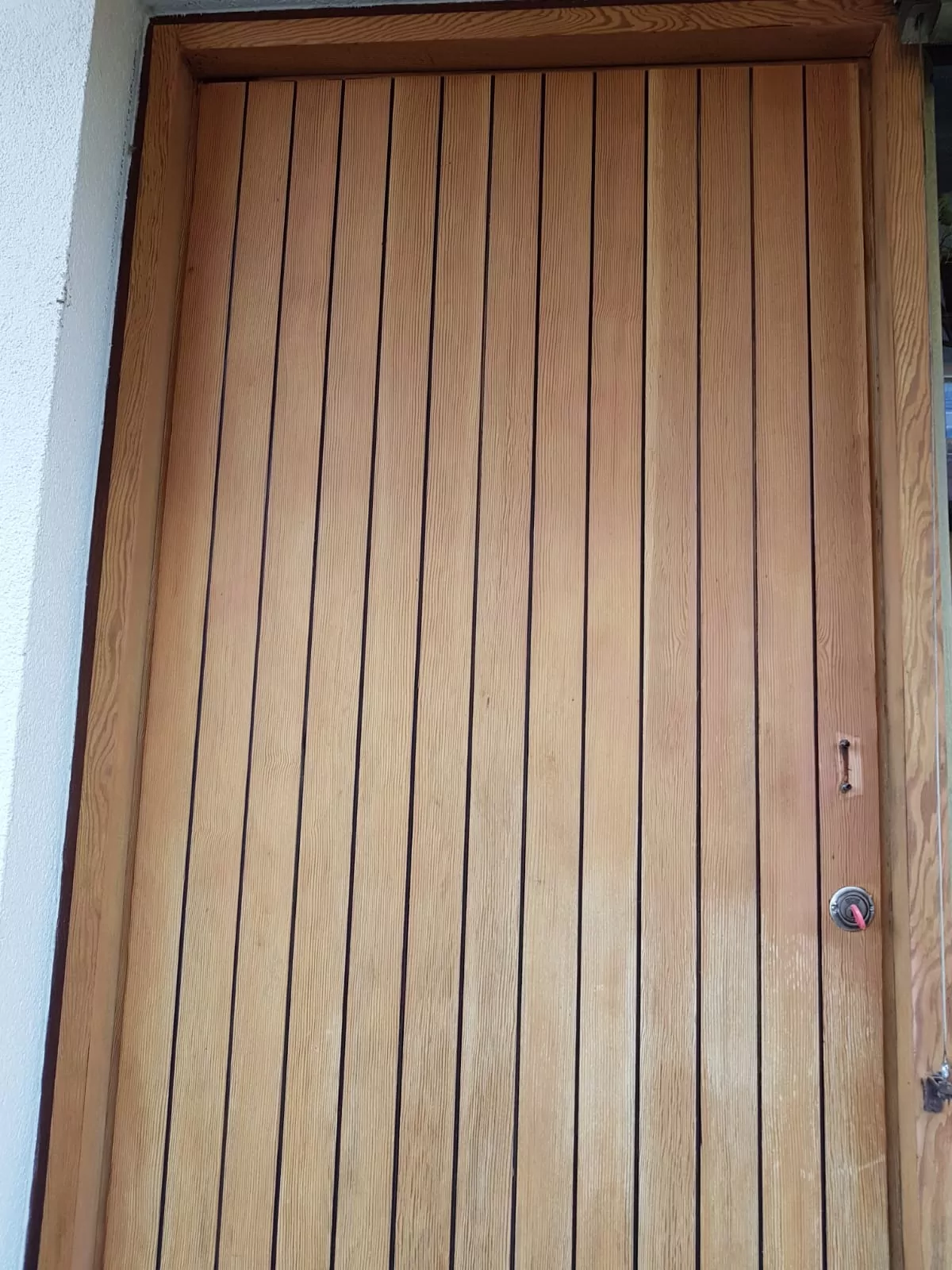 Eine geschlossene Holztür mit vertikalen Brettern, die jeweils durch dünne dunkle Linien voneinander getrennt sind. Auf der rechten Seite nahe der Türkante befindet sich ein runder Türknauf aus Metall. Der Türrahmen ist schlicht gehalten und grenzt an eine helle Wand.