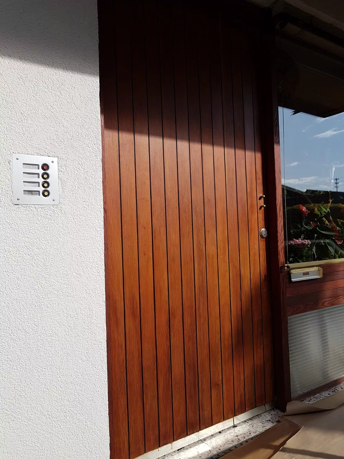 Eine Holztür mit vertikalen Paneelen ist geschlossen. Sie grenzt an eine weiße Wand mit einer Sprechanlage mit drei Tasten. Sonnenlicht wirft Schatten auf die Tür und rechts ist ein Teil eines Fensters mit Blick auf Pflanzen zu sehen.