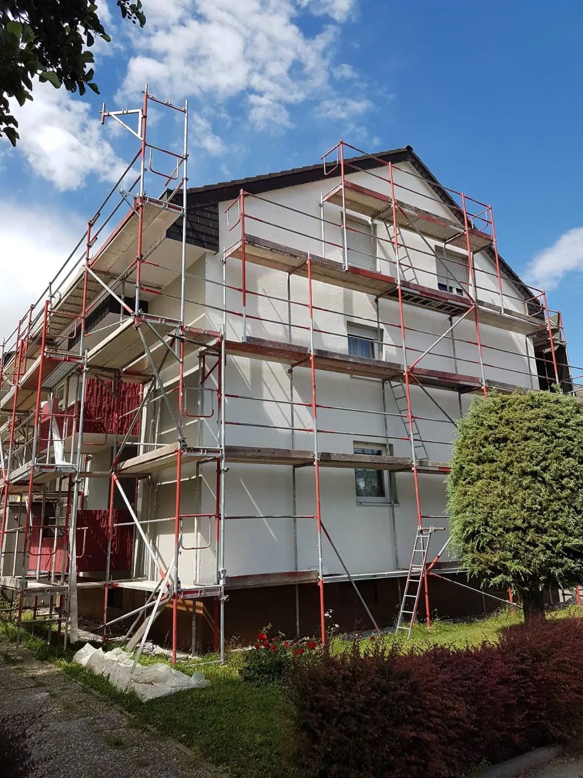 Ein mehrstöckiges Haus ist von roten und silbernen Bau- oder Renovierungsgerüsten umgeben. Der Himmel ist teilweise bewölkt und um das Gebäude herum gibt es Grün, darunter einen gestutzten Busch und kleine Pflanzen.