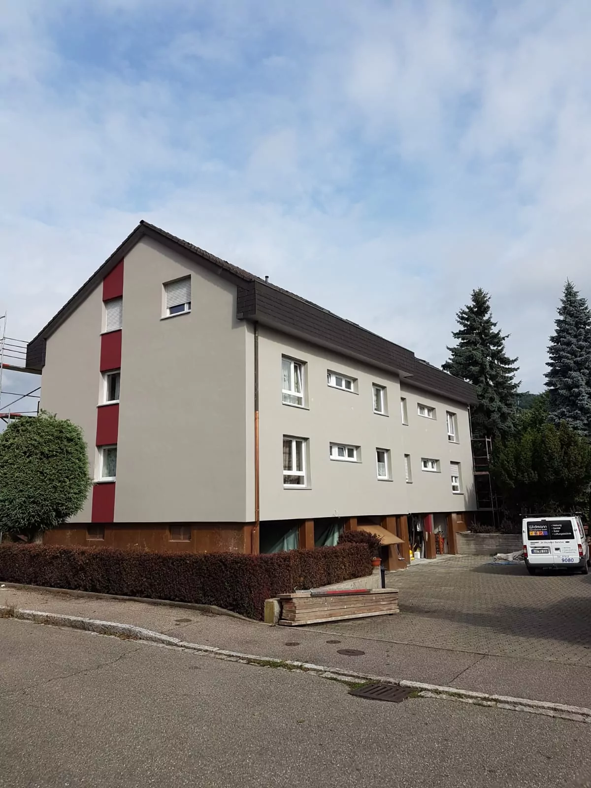 Ein dreistöckiges Wohngebäude mit schrägem Dach und beigen und roten Akzenten. Das Gebäude ist von Bäumen und Büschen umgeben. Auf dem gepflasterten Platz davor parkt ein weißer Lieferwagen. Der Himmel ist teilweise bewölkt.