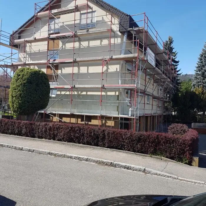 Ein dreistöckiges Haus wird gerade renoviert, umgeben von einem roten Gerüst. Das Gebäude hat eine beige Fassade mit vernagelten Fenstern. Im Vordergrund sind eine ordentlich geschnittene Hecke und ein Baum zu sehen, außerdem ist ein Teil eines geparkten Autos zu sehen.