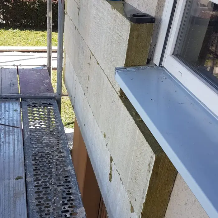 An einem Gebäude ist ein Gerüst aufgebaut, an dem Dämmplatten an einer Außenwand angebracht werden. Auf dem Fensterbrett stehen eine blaue Kiste und ein Werkzeug, darunter sieht man Gras und Gehwege.