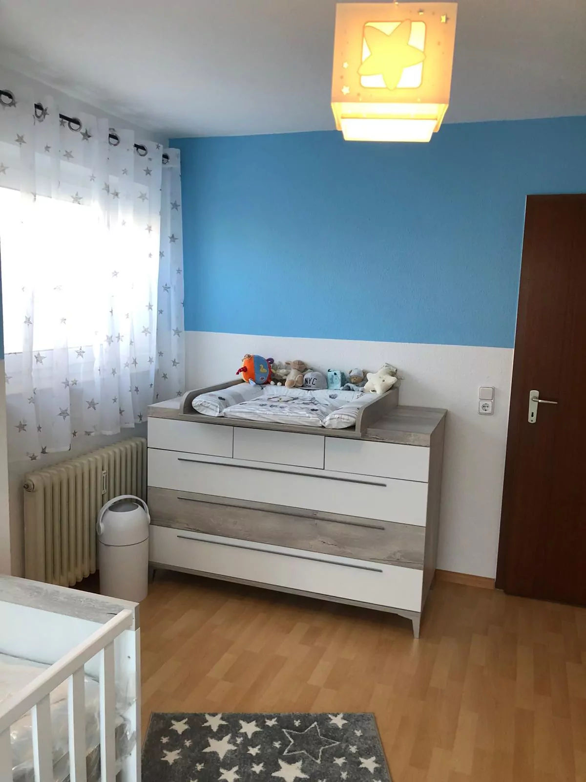 Ein ordentlich organisiertes Kinderzimmer mit einer blauen Akzentwand verfügt über einen Wickeltisch mit Plüschtieren, ein Kinderbett mit weißer Bettwäsche, einen Teppich mit Sternenmuster und einen Vorhang mit Sternenmotiven. Eine quadratische Deckenleuchte erhellt den Raum.