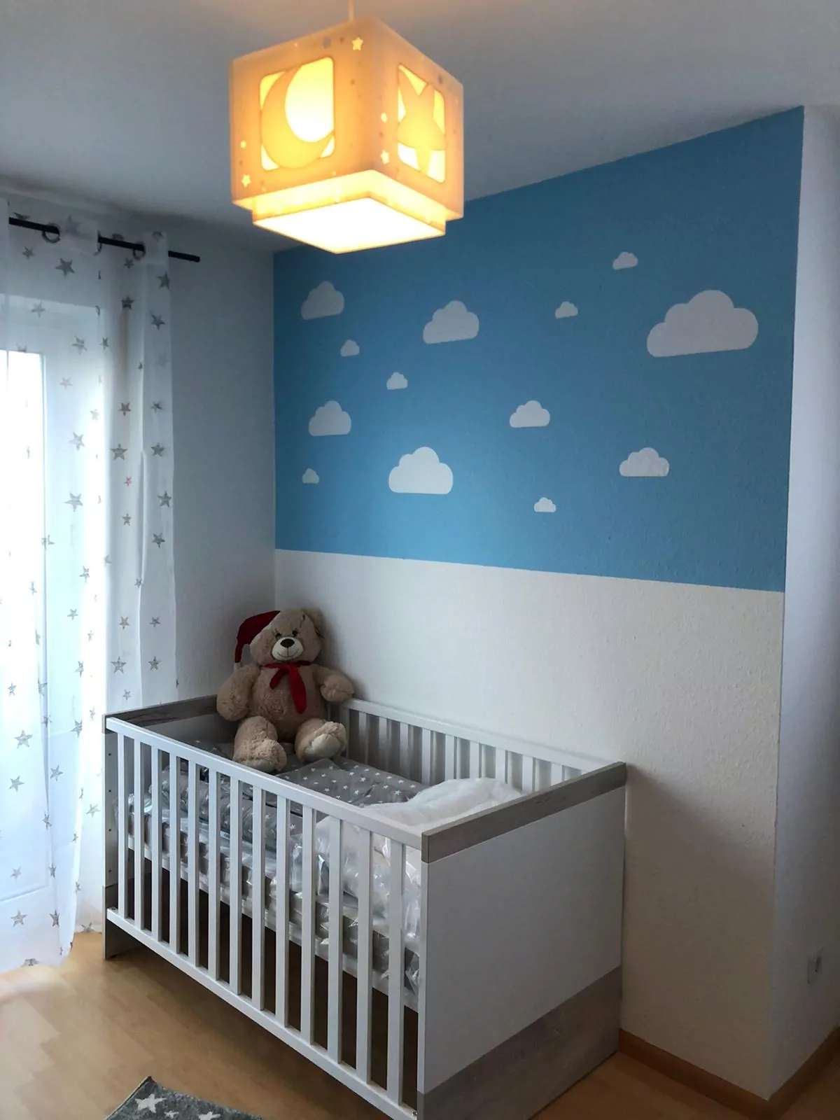 Ein Kinderzimmer mit einem weißen Kinderbett neben einer blauen Wand, die mit Wolkenformen dekoriert ist. Im Kinderbett sitzt ein Teddybär. Am Fenster hängt ein Vorhang mit Sternen- und Mondmuster. An der Decke hängt eine würfelförmige Leuchte mit Mond- und Sternenmuster.