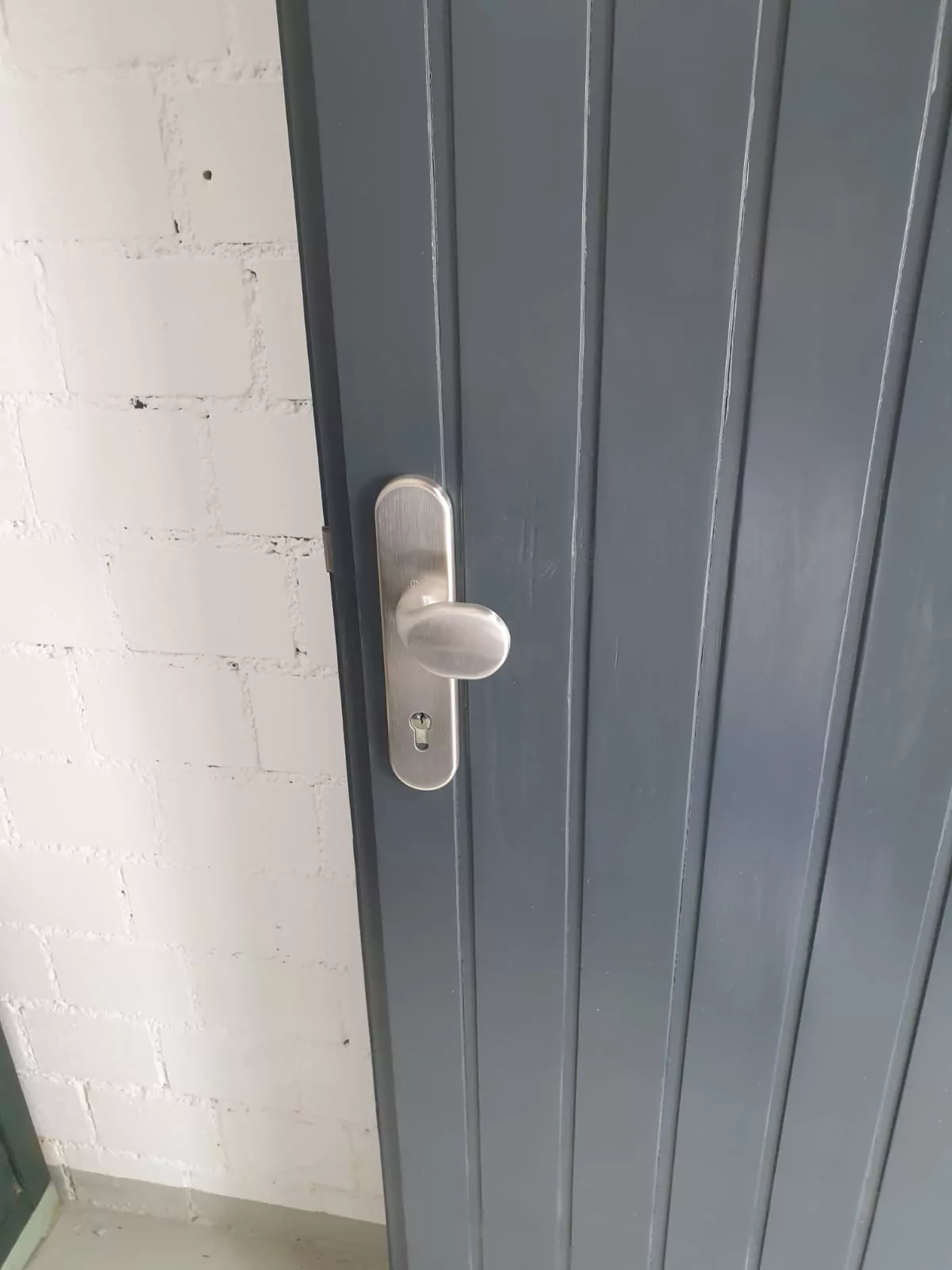 Eine Nahaufnahme eines silbernen Türgriffs mit Schlüsselloch an einer dunkelgrauen, vertikal getäfelten Tür. Die Tür befindet sich neben einer weißen Ziegelwand.