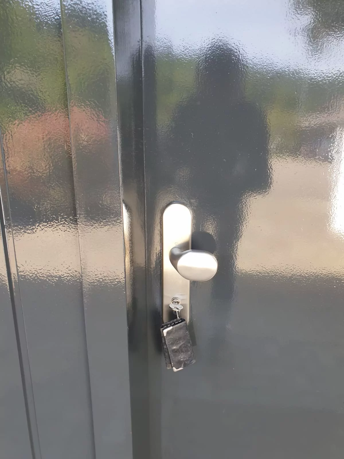 Eine Nahaufnahme einer glänzenden dunklen Tür mit einem silbernen Griff und einem im Schloss steckenden Schlüssel. Auf der Oberfläche der Tür ist eine verschwommene Spiegelung einer Person sichtbar.