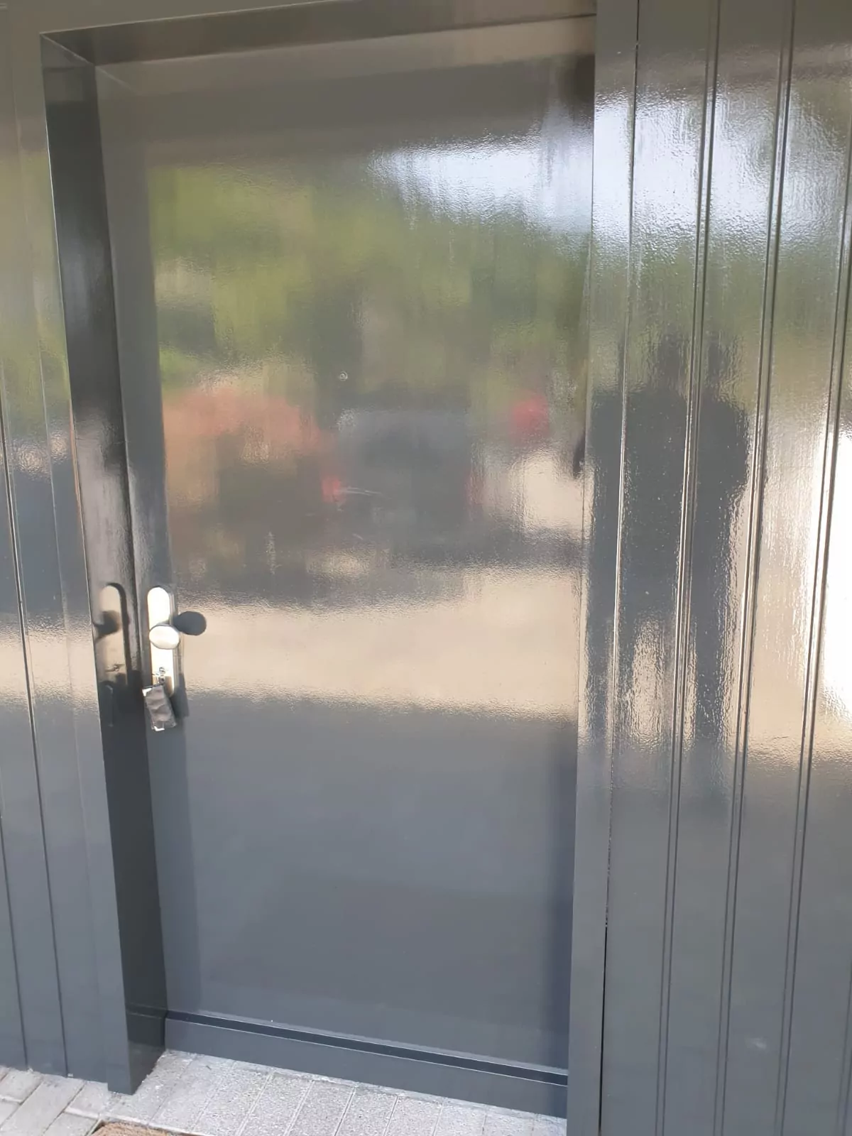 Eine reflektierende, metallische Tür mit elegantem Design, die teilweise die Außenumgebung reflektiert, einschließlich verschwommener Formen und Farben. Die Tür hat einen modernen Griff und ist in eine Struktur mit passender Verkleidung eingelassen.