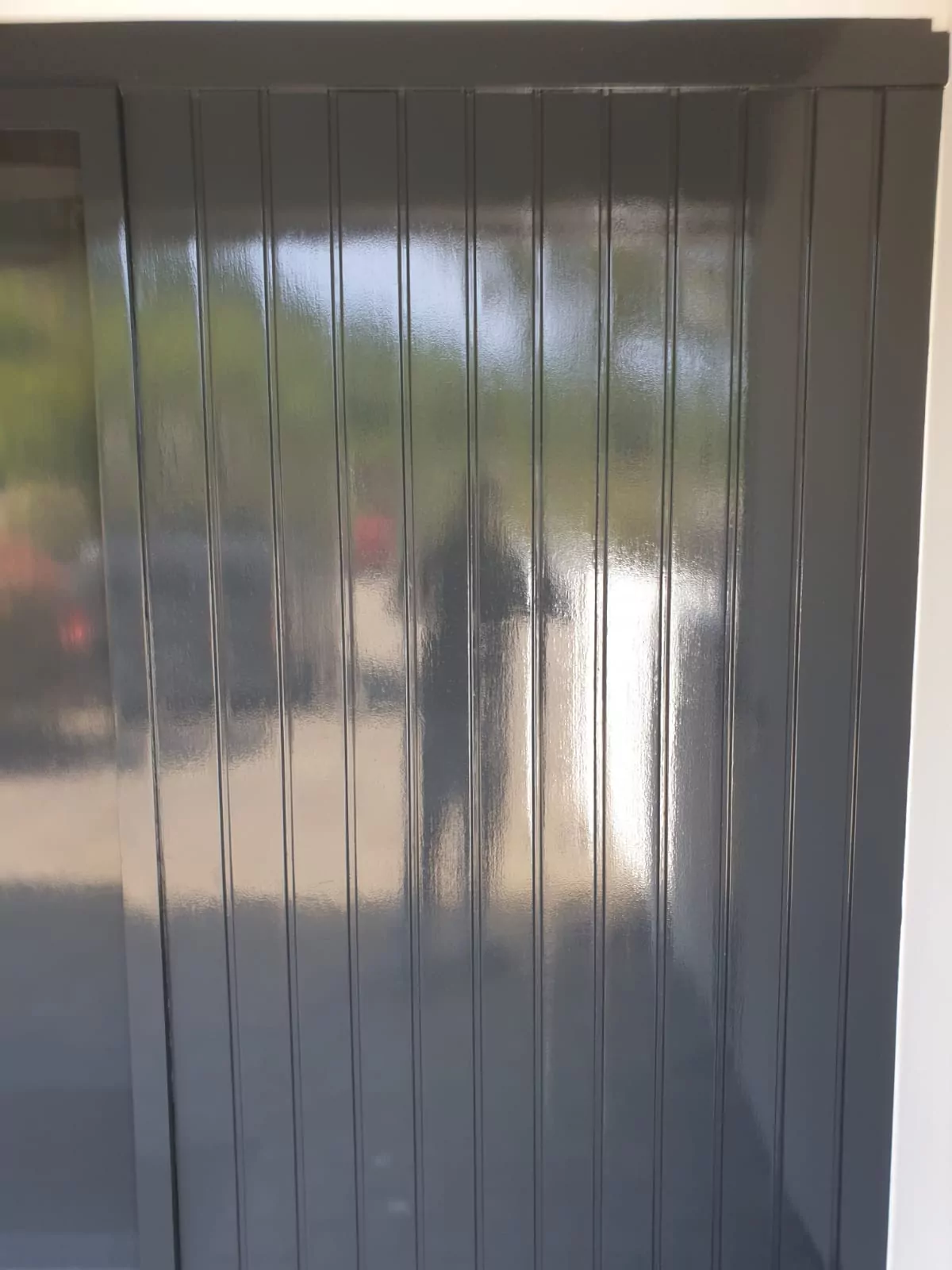 Spiegelung einer Person, die ein Foto in einem glänzenden, dunkel getäfelten Garagentor macht. Die Außenszene, darunter etwas Grün und ein rotes Auto, ist in der Spiegelung sichtbar.