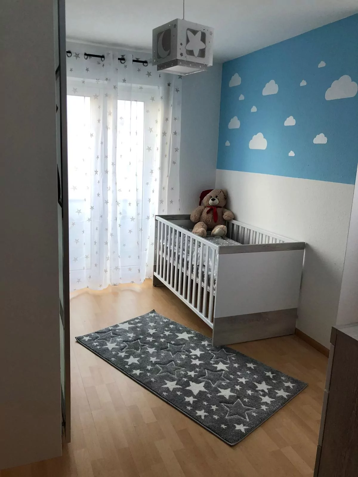 Ein kleines Kinderzimmer mit einem Kinderbett mit einem Teddybären, einer blauen Wand mit Wolken- und Monddekorationen und einem grauen Teppich mit Sternen auf einem Holzboden. Weiße Vorhänge mit Sternendrucken bedecken ein Fenster und darüber hängt eine Lampe mit Stern- und Mondformen.