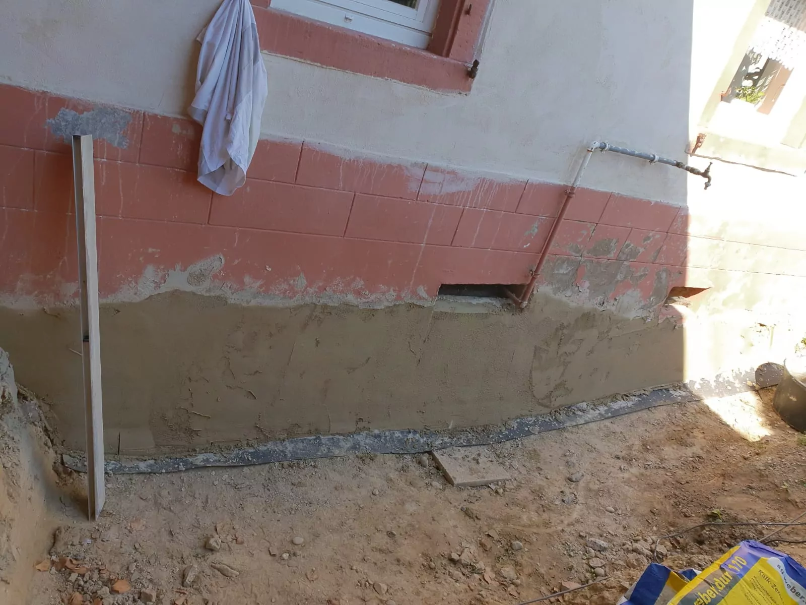 Ein Abschnitt einer Außenwand eines Hauses zeigt Reparaturarbeiten. Der untere Teil der Wand ist frisch verputzt, während der obere Teil noch seine ursprüngliche Farbe hat. Ein langes Werkzeug lehnt an der Wand und ein Stück Stoff hängt daneben. Der Boden besteht aus unebenem Schmutz.