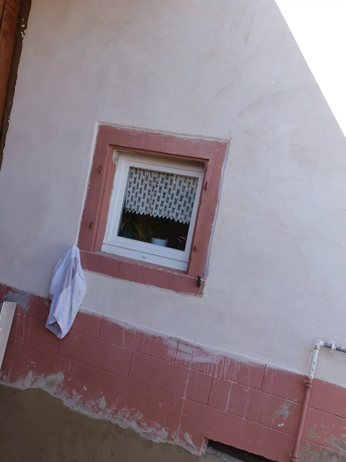 Ein kleines Fenster mit einem Spitzenvorhang in einer Stuckwand, eingerahmt von rosa Zierleisten. Links vom Fenster hängt ein weißes Handtuch. In der oberen rechten Ecke des Gebäudes ist ein schräger Dachschatten zu sehen.