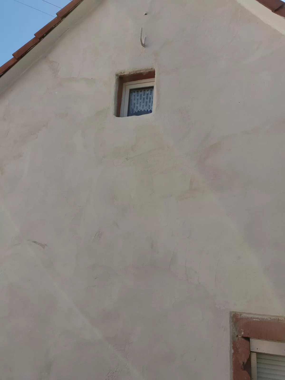 Eine beige Stuckwand mit einem einzigen kleinen quadratischen Fenster oben, das teilweise von einem Spitzenvorhang bedeckt ist. Das Fenster hat einen braunen Rahmen und über der Dachlinie ist der Himmel sichtbar.