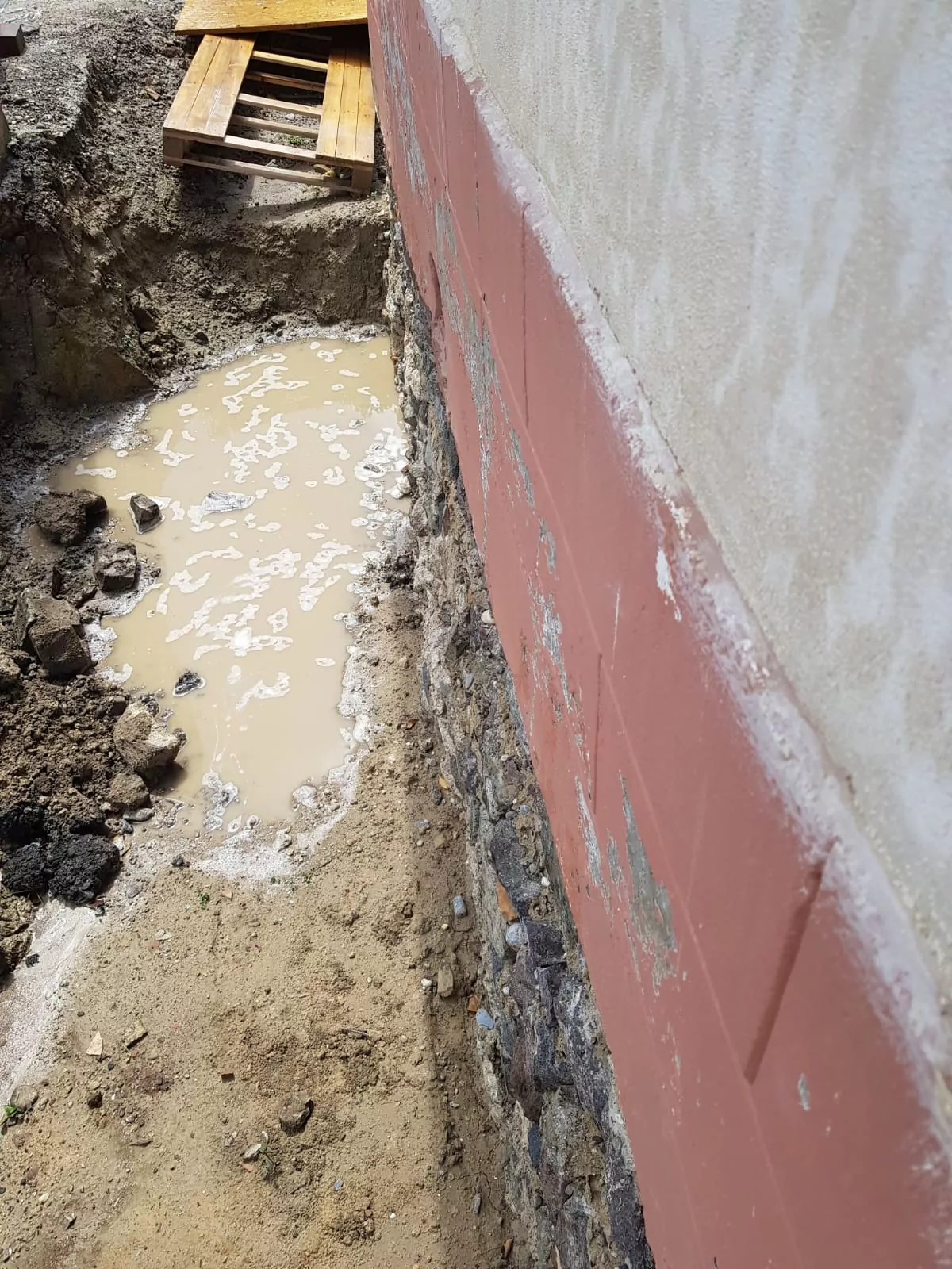 Ausgrabungsfläche neben einem Gebäude mit freiliegendem Fundament aus Steinen und Zement. Der Boden ist schlammig und enthält eine kleine Wasserpfütze. Im Hintergrund ist teilweise eine Holzpalette zu sehen.