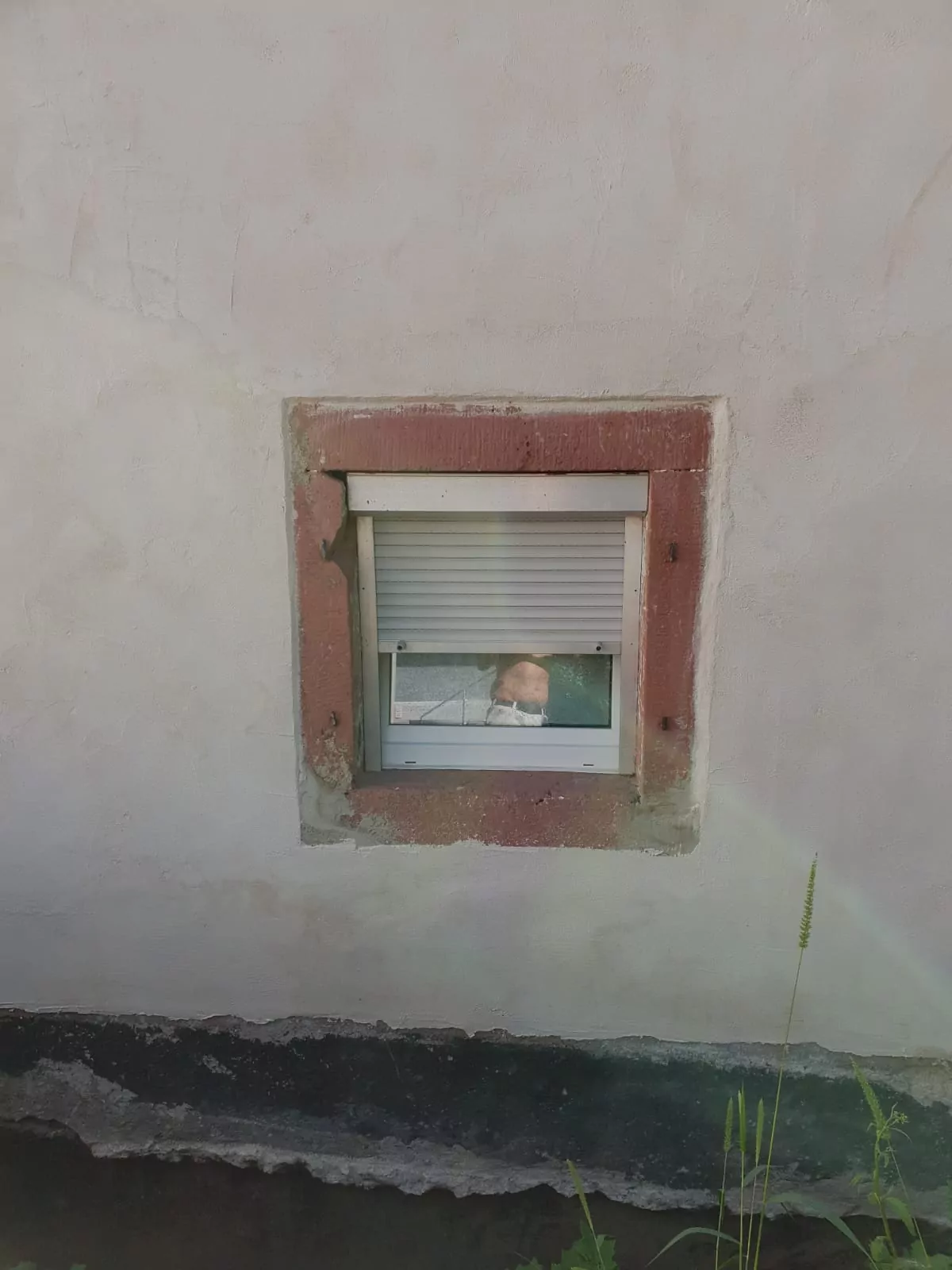 In einer verwitterten Wand befindet sich ein kleines Fenster mit einer teilweise geschlossenen weißen Jalousie. Durch das Fenster sieht man ein Baby, das nach draußen blickt. Die Wand weist Alterserscheinungen mit Verfärbungen und Rissen auf. Auf der Wand ist ein schwacher Regenbogen zu sehen.