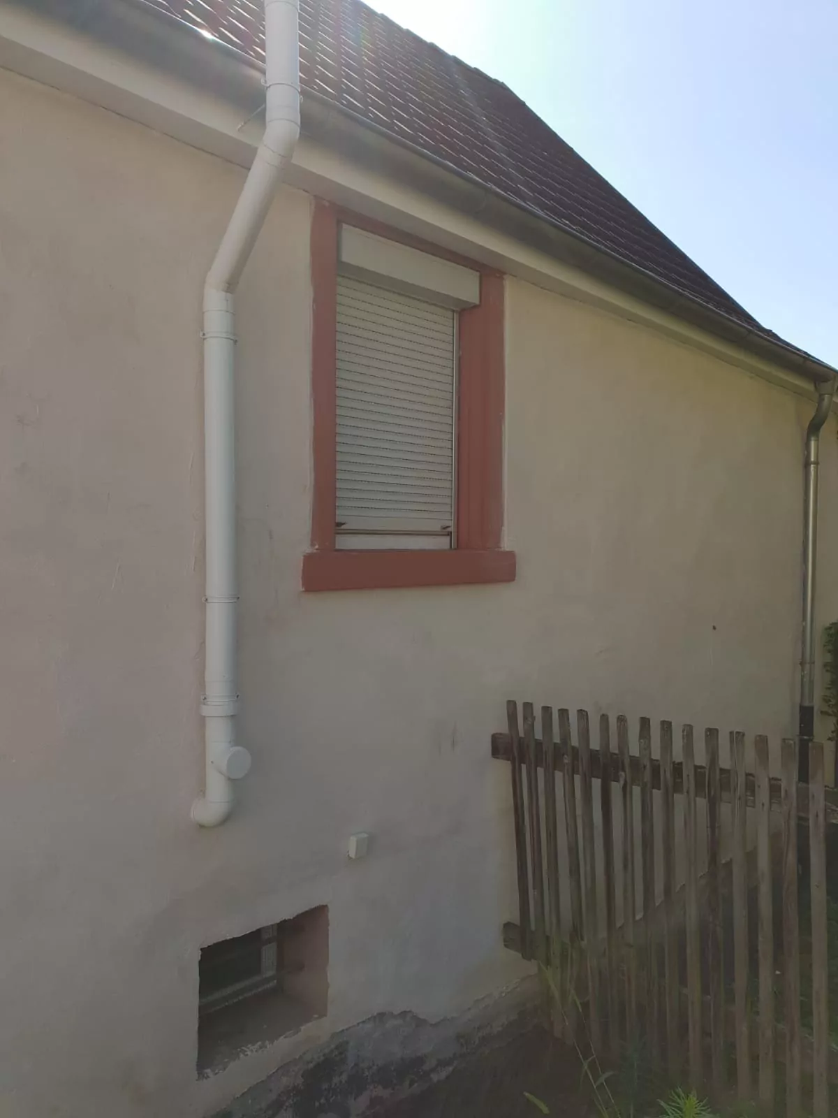 Ein kleines, beiges Haus mit einem einzigen Fenster hat einen teilweise geschlossenen weißen Rollladen. Ein weißes Regenfallrohr verläuft entlang der Wand. Im Vordergrund ist ein Holzzaun zu sehen. Der Himmel ist klar und sonnig.