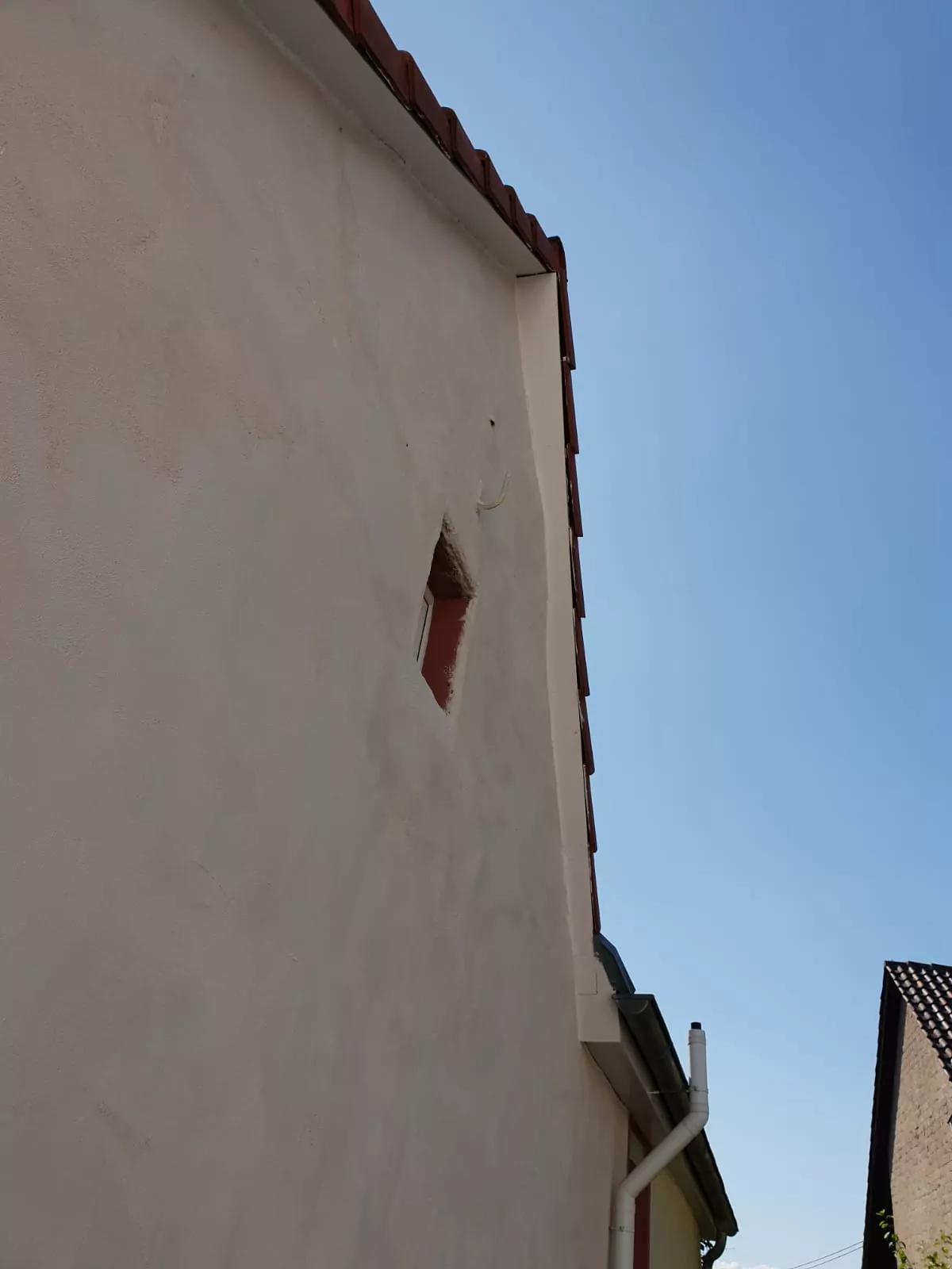 Eine beige Stuckwand mit einer kleinen rechteckigen Öffnung in Dachnähe und einem rot gefliesten Rand. An der Seite verläuft eine weiße Dachrinne. Der Himmel ist klar und blau.