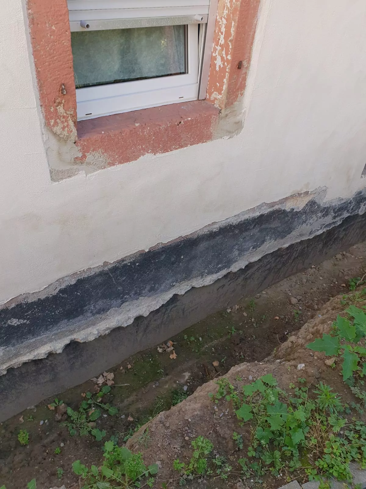 Eine Gebäudewand mit sichtbaren Putzschäden, die freiliegende Schichten zeigen. Unter dem Fundament befindet sich ein Streifen dunkler Feuchtigkeitsbarriere und Erde. Der Bereich ist ungepflegt, in der umgebenden Erde wachsen Grasflecken und Unkraut.