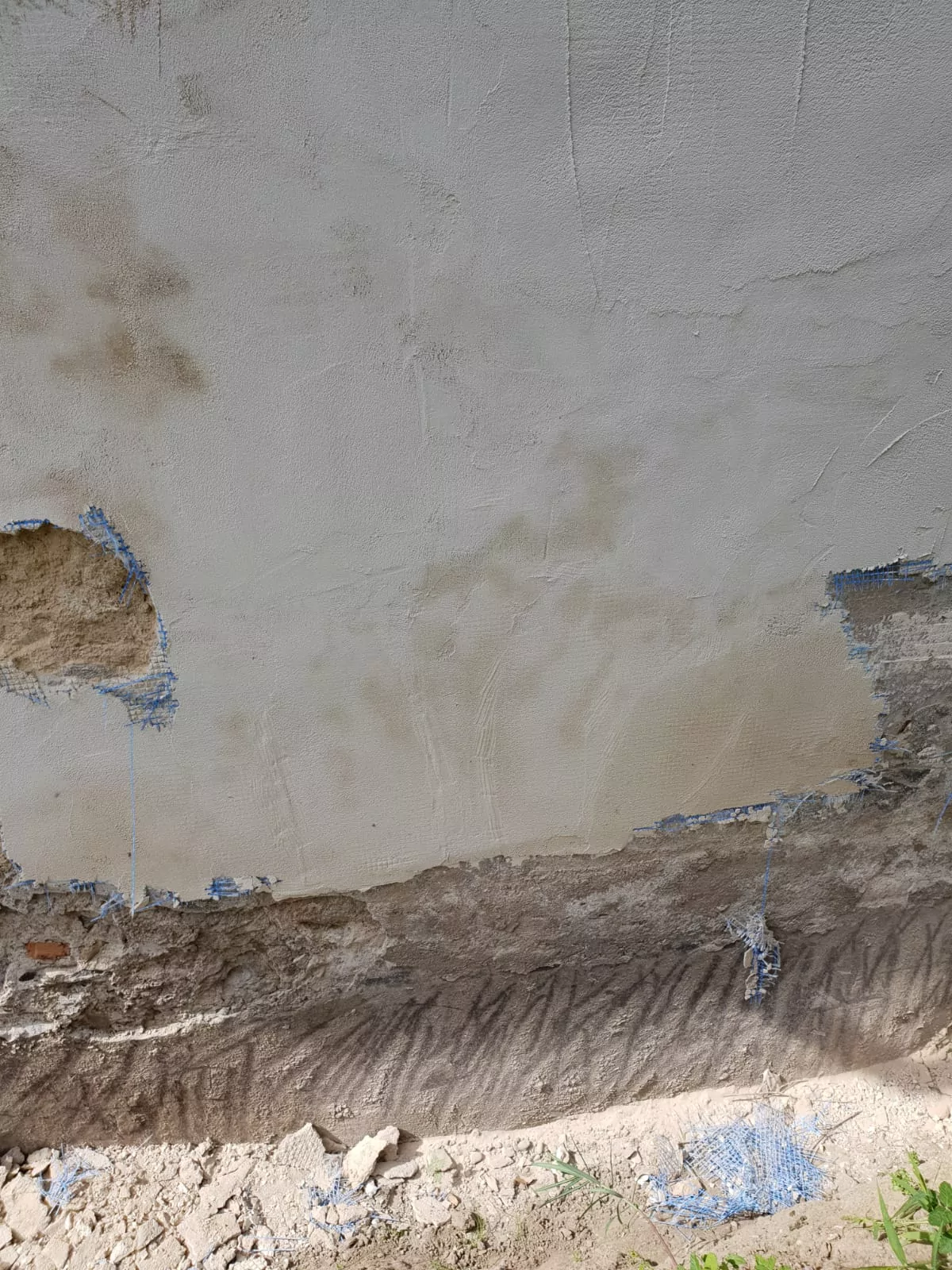 Eine Wand, deren Putz sich teilweise ablöst, wodurch die raue, verwitterte Oberfläche darunter sichtbar wird. Der Boden darunter ist sandig, am Rand ist etwas Grün zu sehen. Sonnenlicht wirft Schatten auf den unteren Teil der Wand.