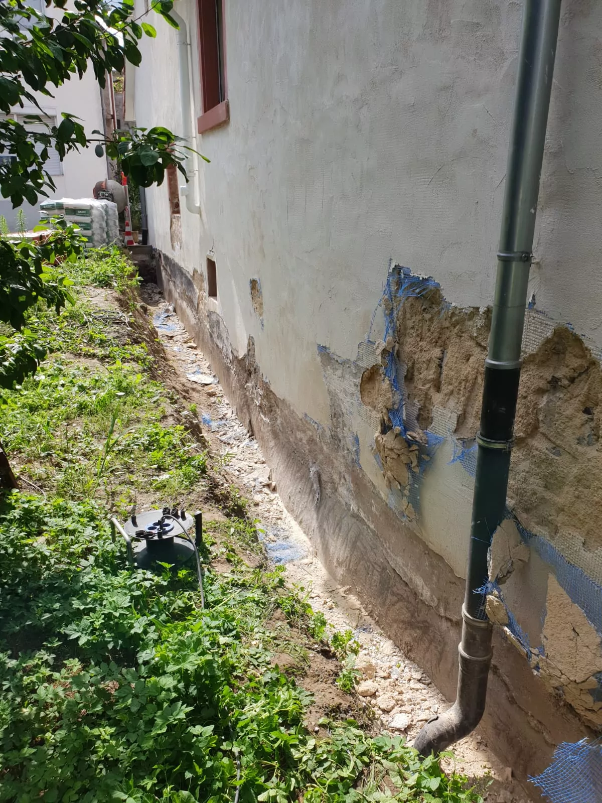 Eine Hausfassade mit sichtbaren Schäden am Putz der unteren Wand. Der Boden ist uneben und weist einige kahle Stellen auf. Ein Regenfallrohr verläuft entlang der Wand. Auf der linken Seite ist Grün zu sehen, im Hintergrund sind Säcke und ein Fenster zu sehen.